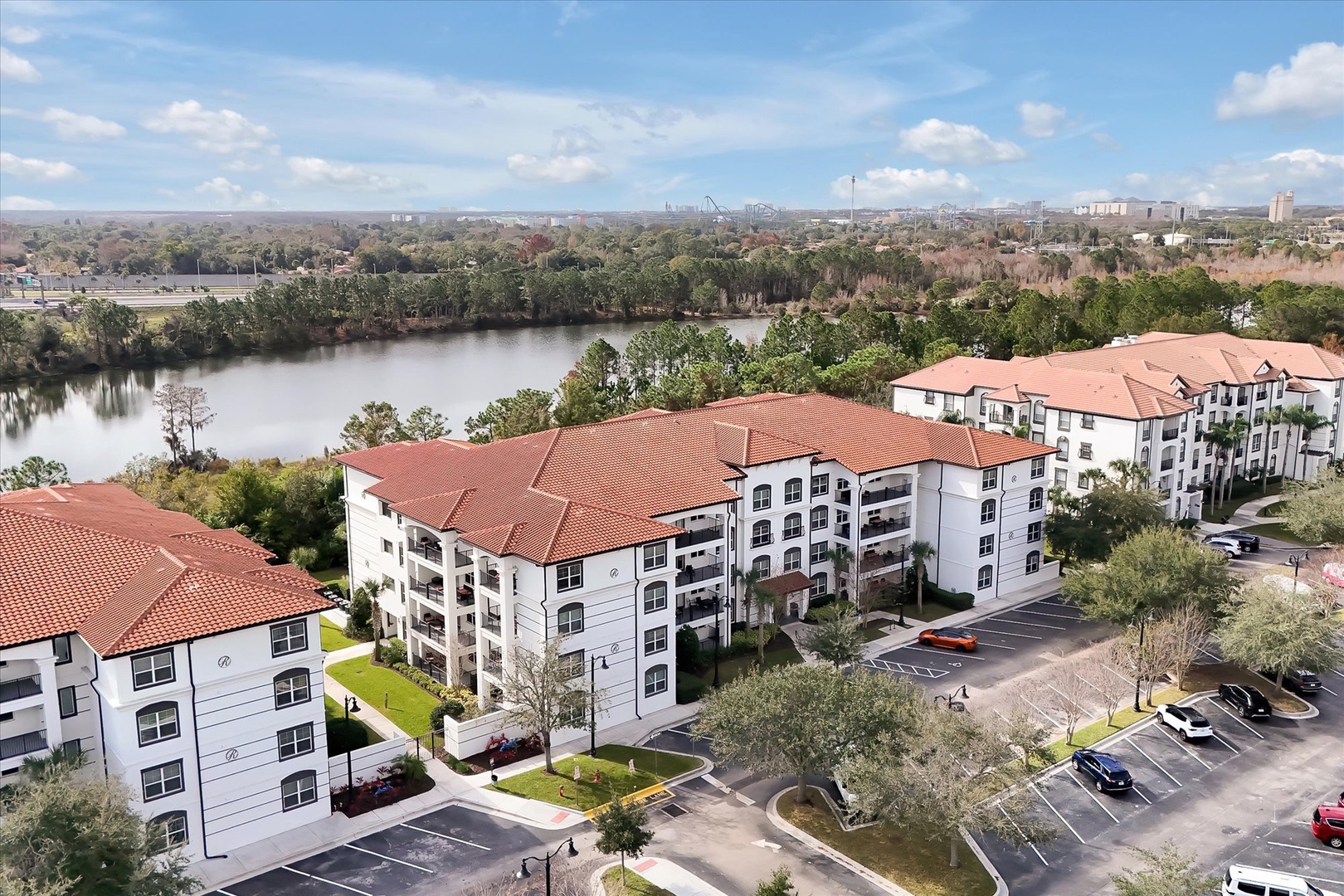 Waterfront property complex featuring Mediterranean-style buildings with red tile roofs overlooking a peaceful lake and urban skyline.