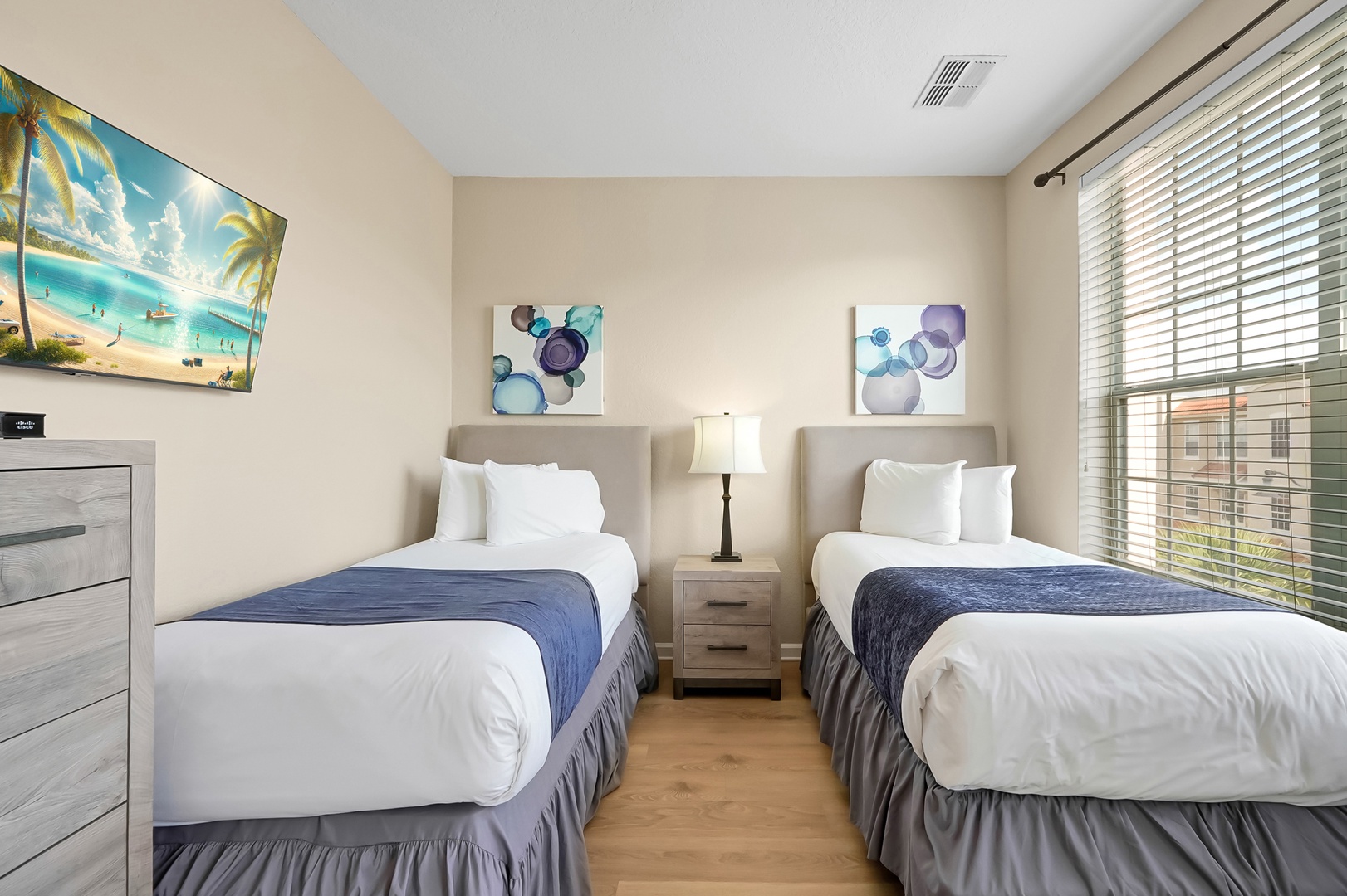 Settle into your peaceful twin bedroom with crisp linens, tropical artwork, and warm natural light streaming through the blinds.