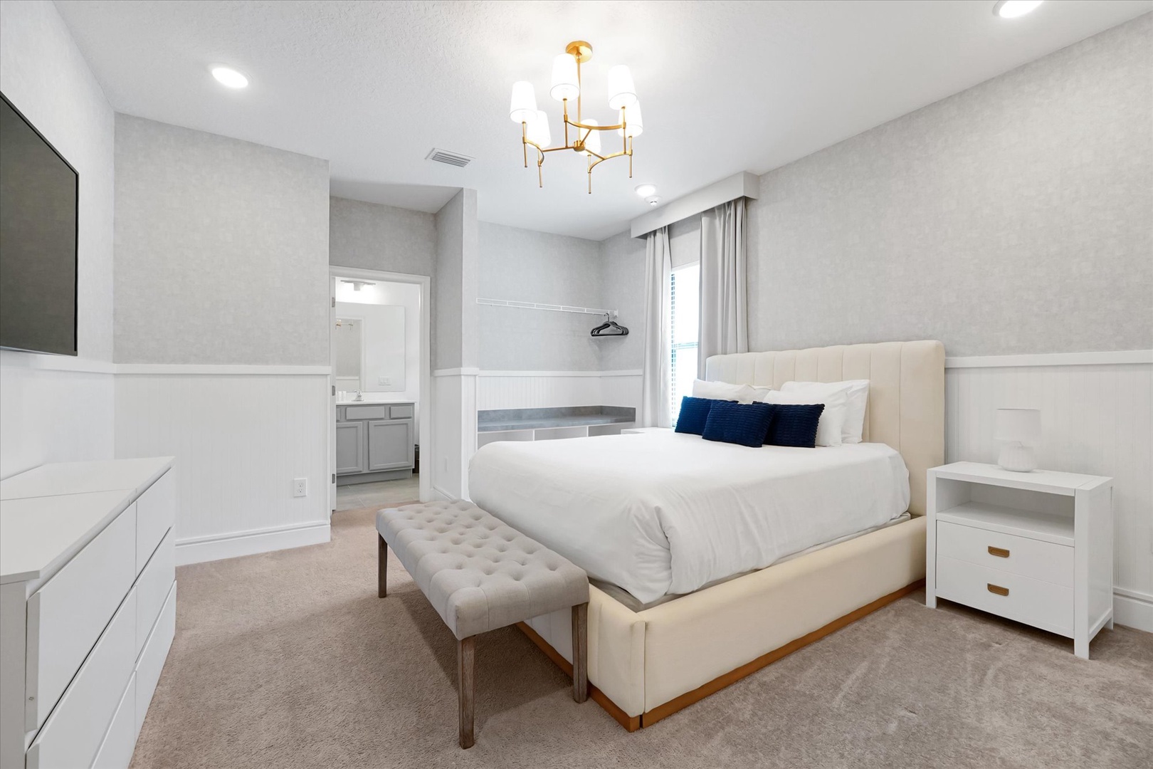Your serene bedroom retreat features a plush upholstered bed with crisp linens, elegant chandelier overhead, and an ensuite bathroom just steps away. Relax in this peaceful space designed for your comfort.