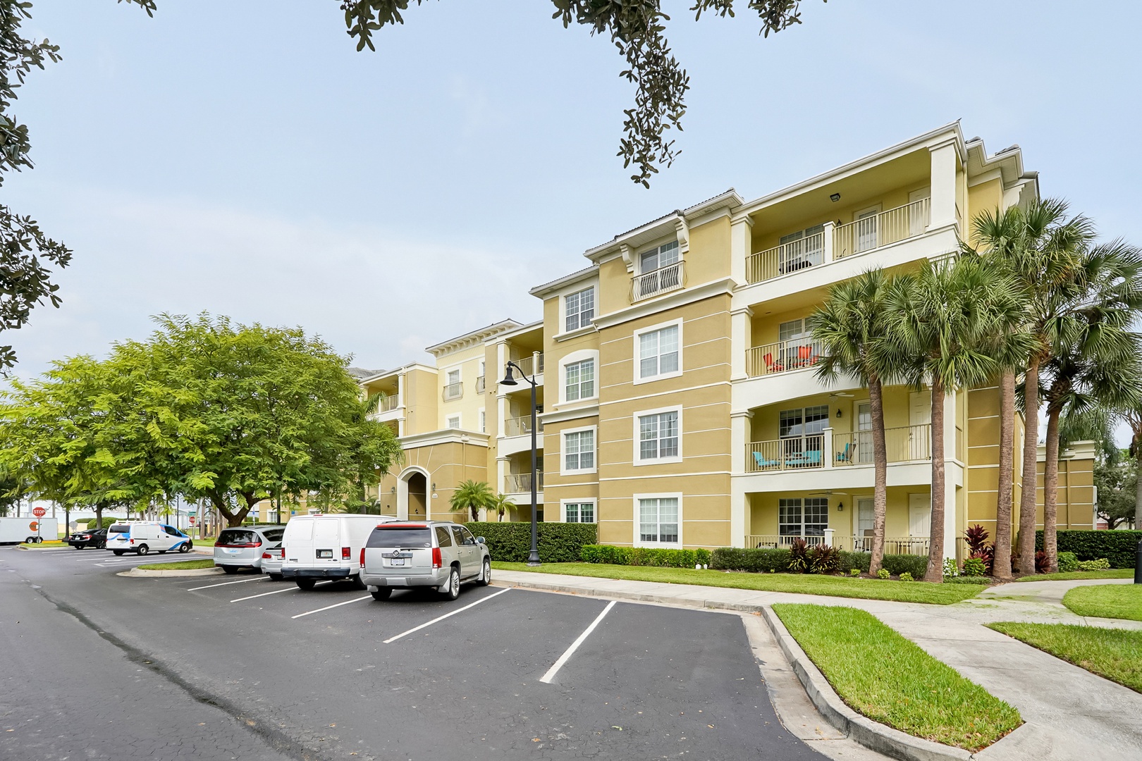 The property building features a welcoming yellow façade with multiple balconies, surrounded by lush tropical landscaping and convenient parking for guests.