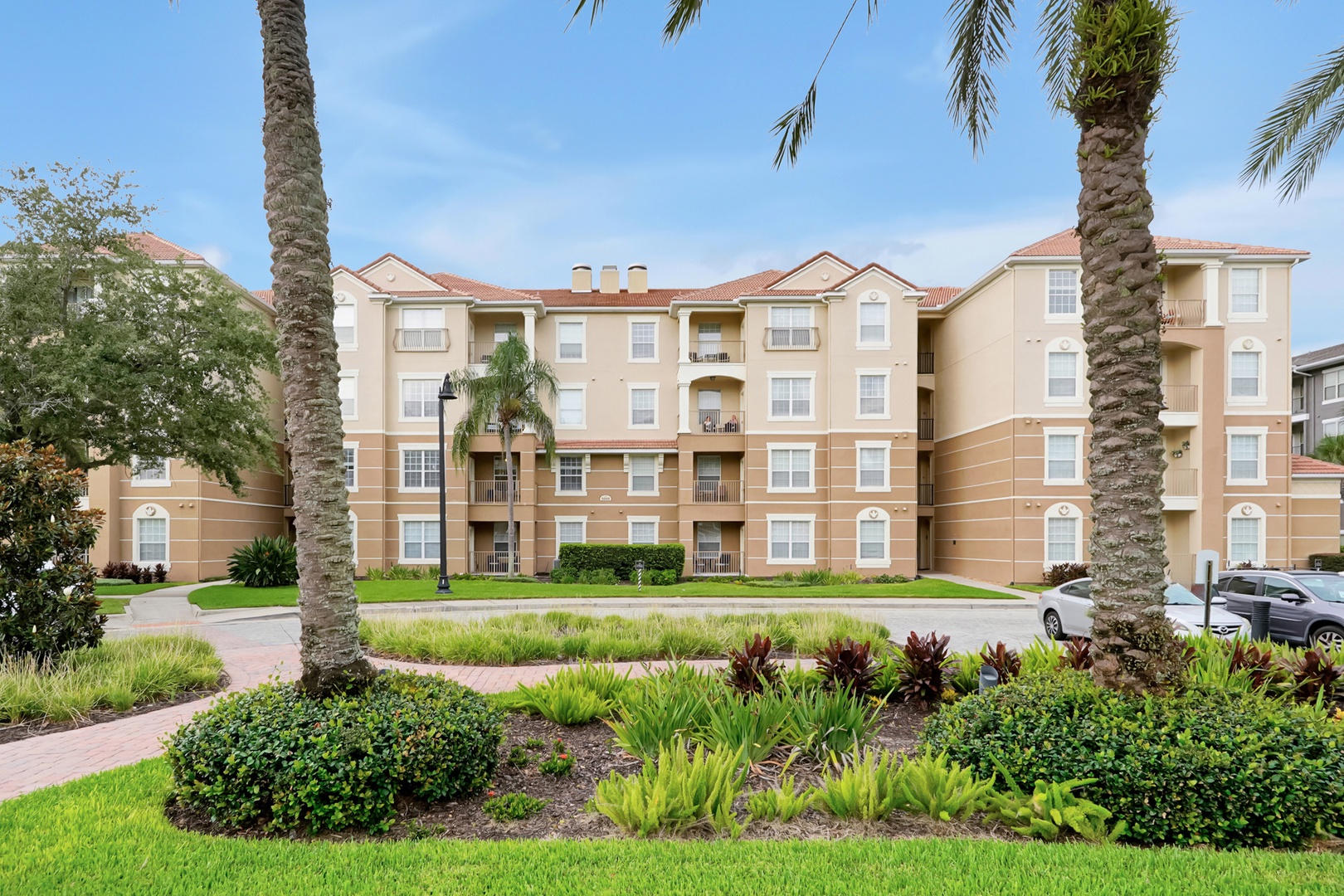 Located in building 5000, right next to the clubhouse. Exterior view of the property building, framed by lush tropical landscaping and mature palm trees, with manicured lawns and greenery throughout the grounds.