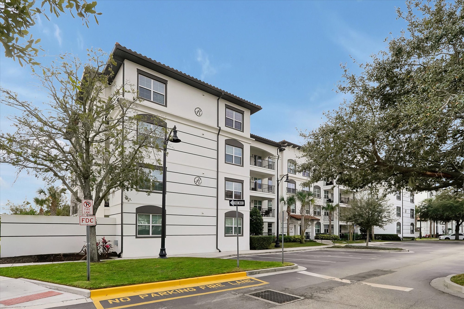 Modern white apartment complex with Mediterranean-style architecture nestled among mature trees on a quiet residential street.