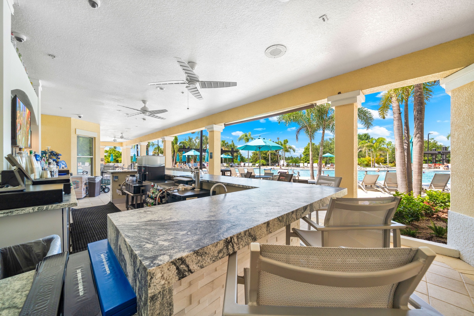 A covered outdoor bar and dining area overlooks the resort pool, framed by swaying palms and sunny blue skies. The perfect spot to relax with a refreshing drink after a day in the Florida sun.