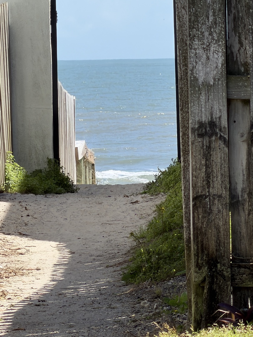 A quiet coastal pathway frames the approach to the nearby beach, offering glimpses of sparkling waves and soft sand just steps from the property.