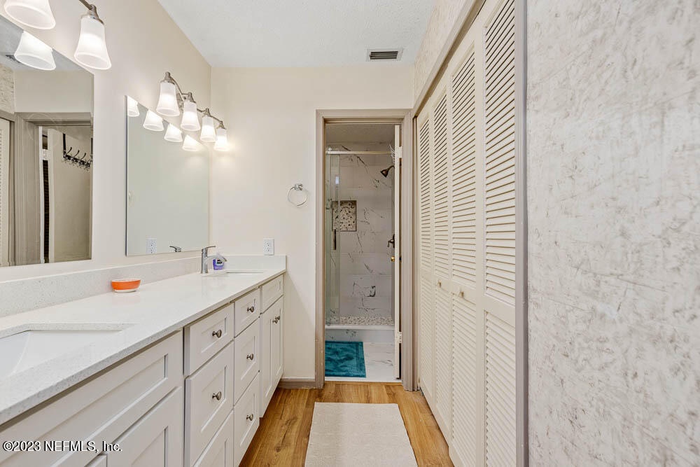 Your spacious bathroom offers gleaming marble surfaces, ample storage with white cabinetry, and bright vanity lighting. A serene walk-in shower awaits just beyond, with a turquoise accent mat to greet your feet.