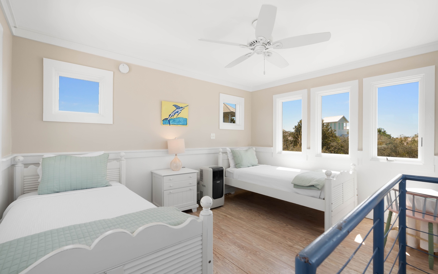 Relax in this bright coastal bedroom with twin beds, ceiling fan, and stunning windows showcasing peaceful neighborhood views.
