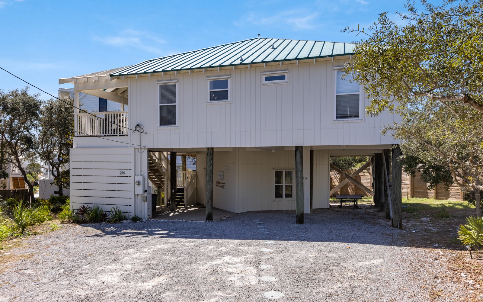 Coastal-style elevated home with metal roof and covered parking, surrounded by mature trees in a peaceful neighborhood setting.