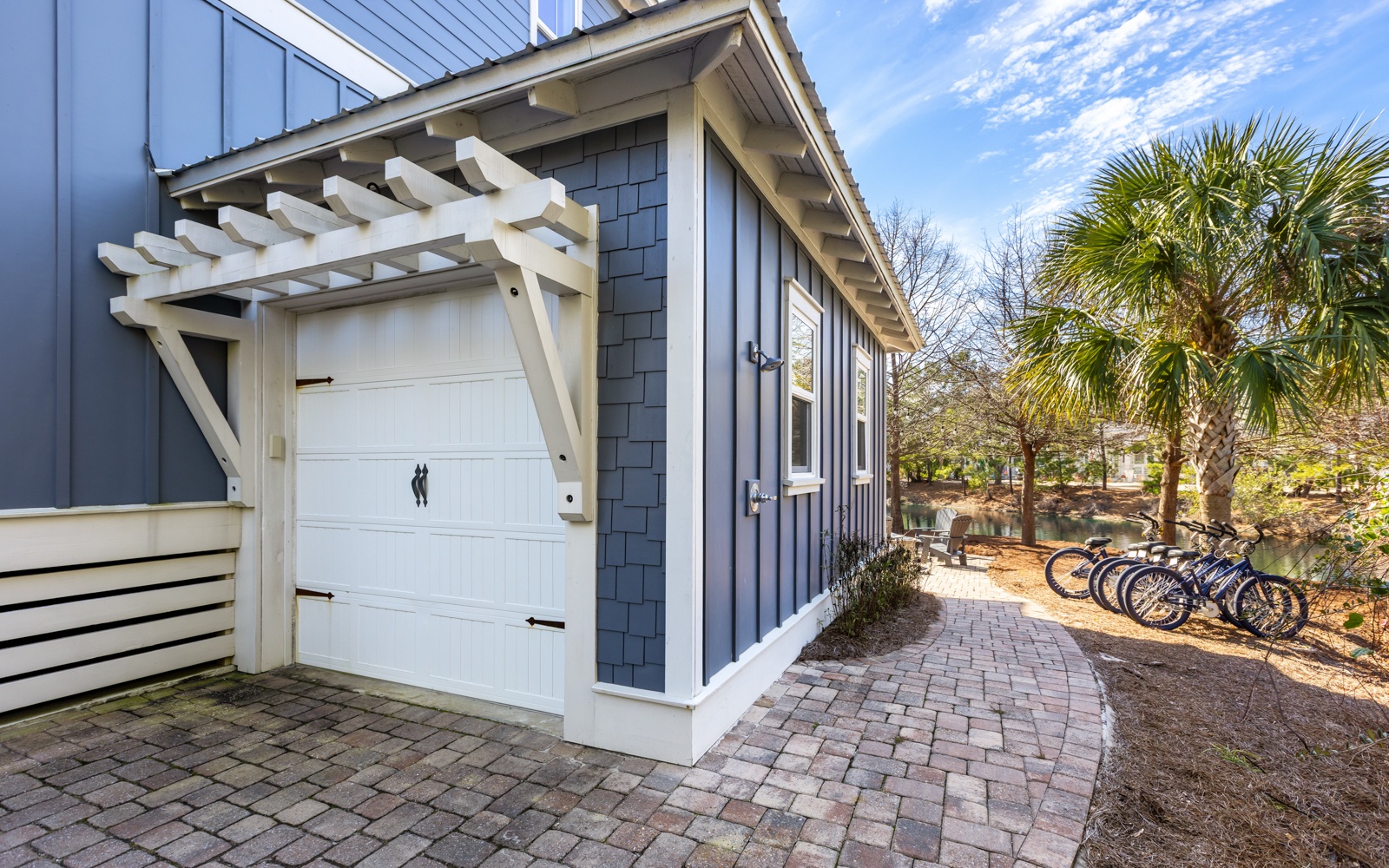 Modern beach house exterior with private garage and bike storage, surrounded by tropical palms and brick walkways.