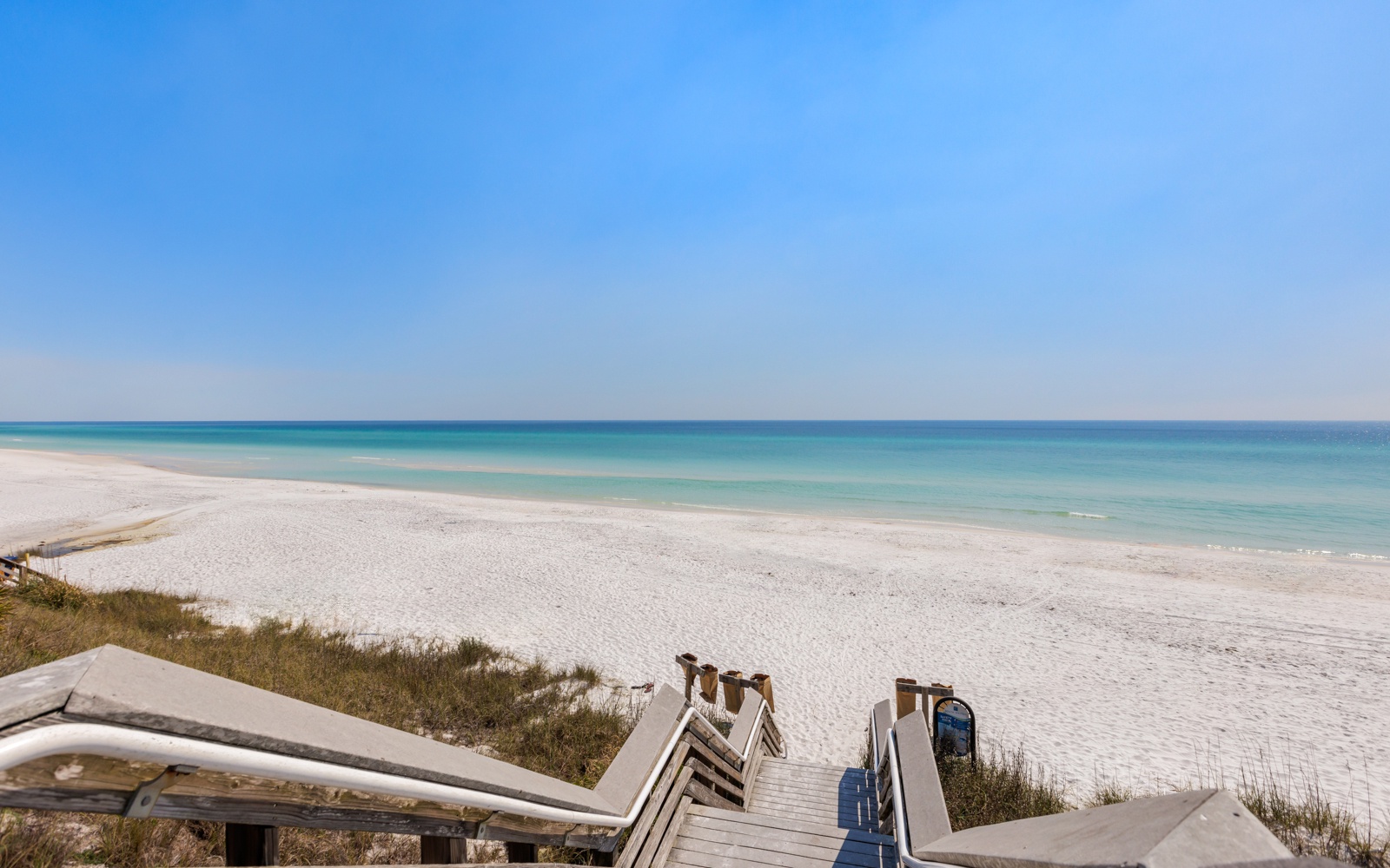 Pristine white sand beach stretches endlessly along turquoise waters under brilliant blue skies, accessible via wooden boardwalk stairs.