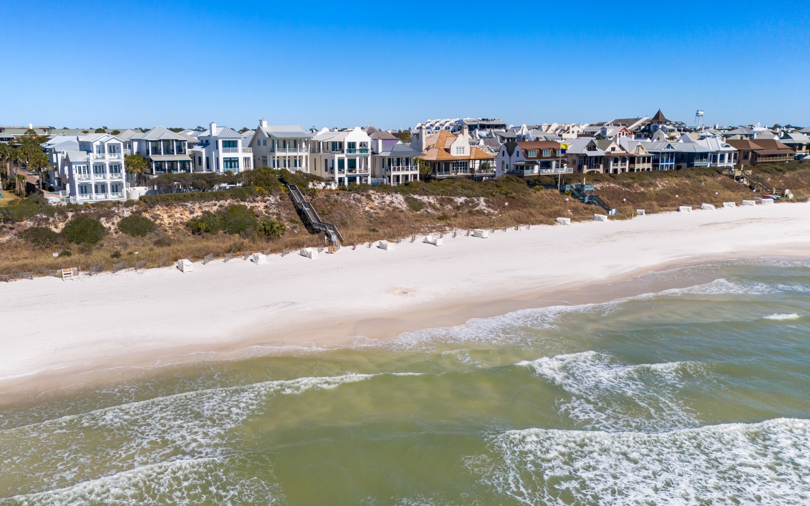 Stunning aerial view of beachfront homes nestled along pristine white sand shores with crystal-clear emerald waters.