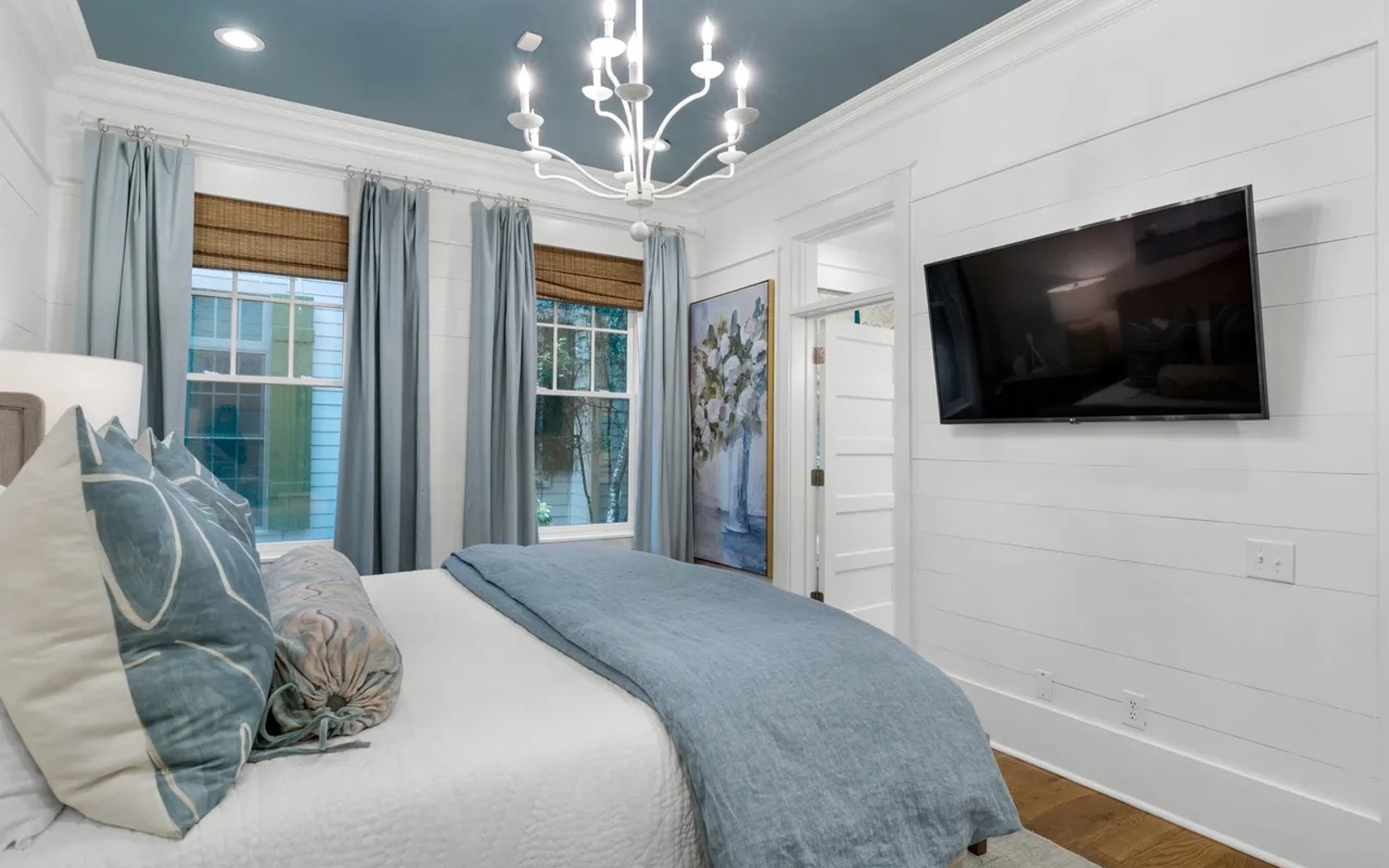 Drift off peacefully in your coastal-inspired bedroom with crisp white shiplap walls, elegant chandelier, and soft blue accents creating serene comfort.