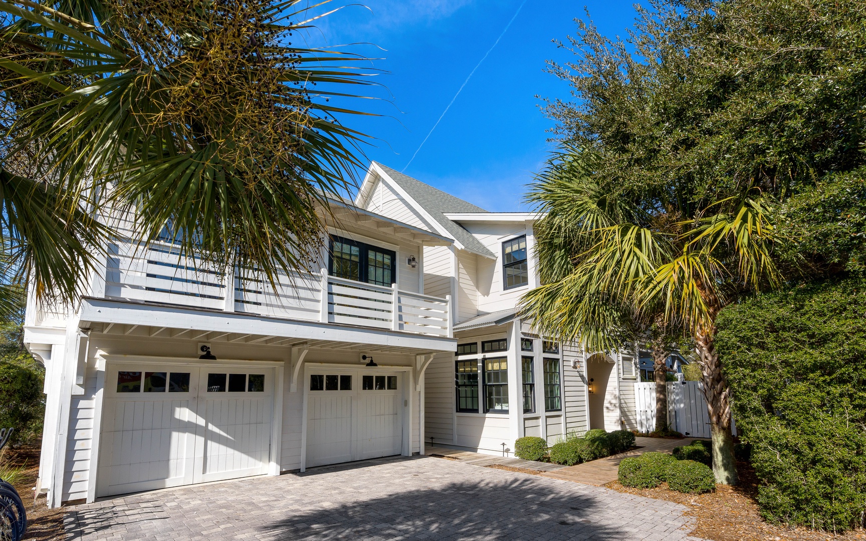 Modern two-story coastal home with elegant white siding and covered parking, nestled among mature palm trees under bright blue skies.