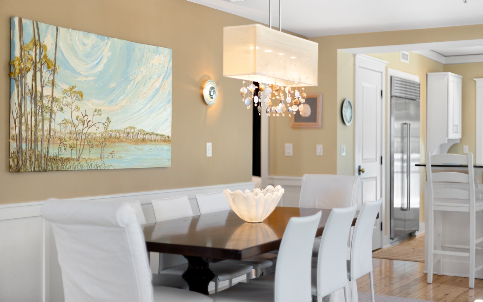 Gather around this elegant dining table for memorable meals, bathed in warm chandelier light with coastal artwork setting the scene.