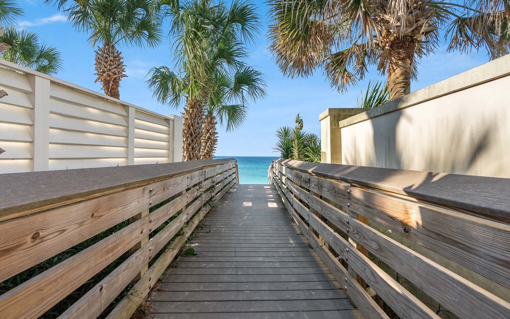 Wooden boardwalk leads through tropical palms to pristine turquoise waters and sandy beach beyond.
