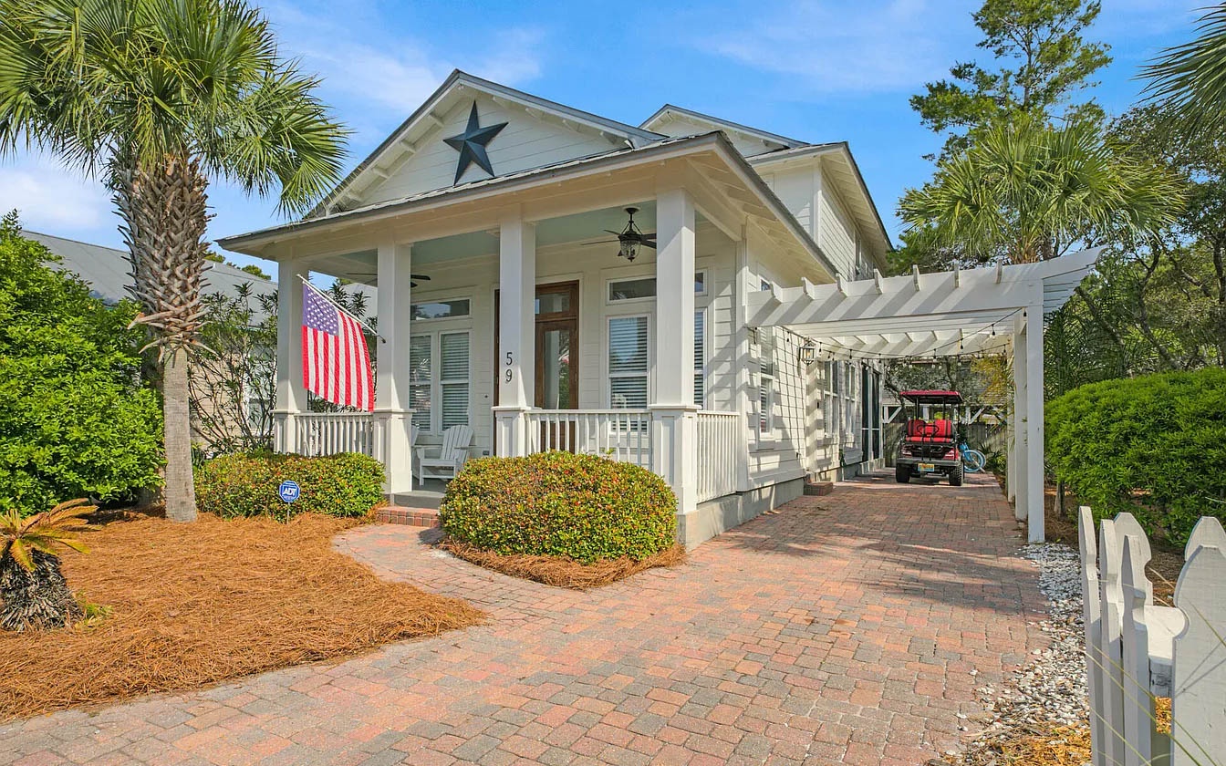Charming coastal cottage with star-adorned facade, palm-lined entrance, and convenient carport parking welcomes guests to this peaceful neighborhood retreat.
