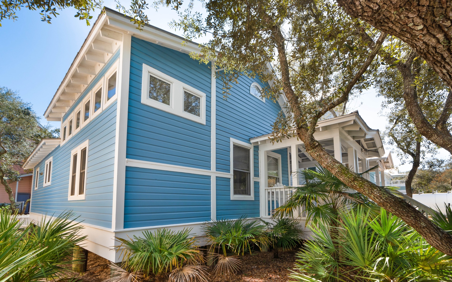 Charming blue coastal cottage surrounded by mature trees and tropical landscaping in a peaceful neighborhood setting.