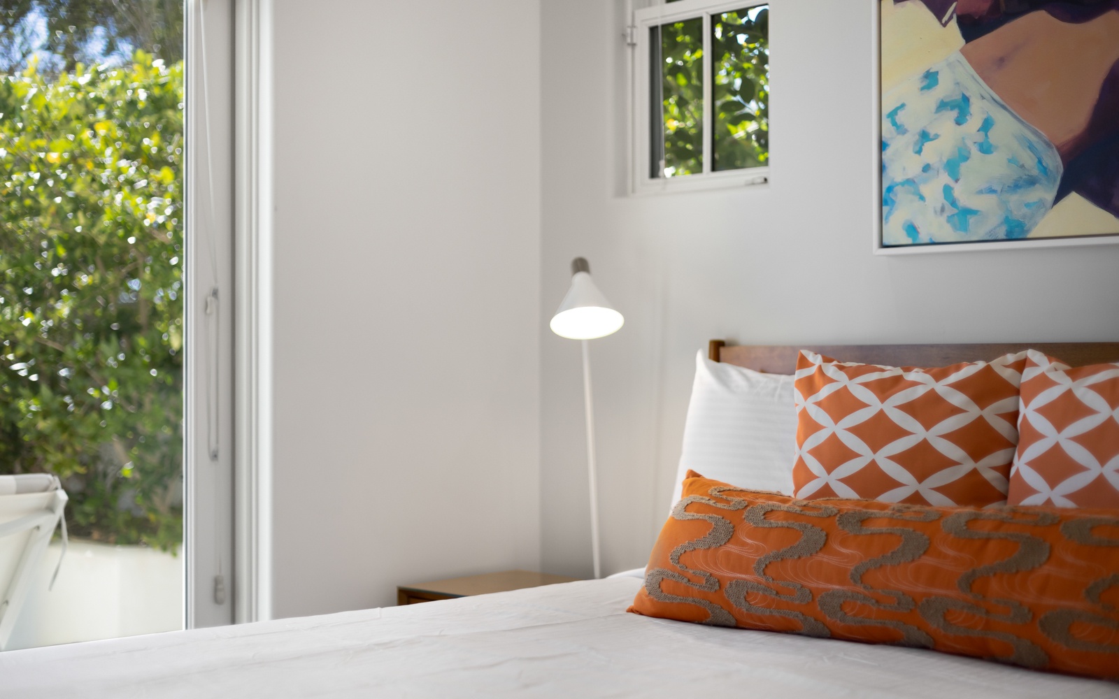 Sink into crisp white linens adorned with vibrant orange pillows, while natural light streams through your windows overlooking lush greenery.