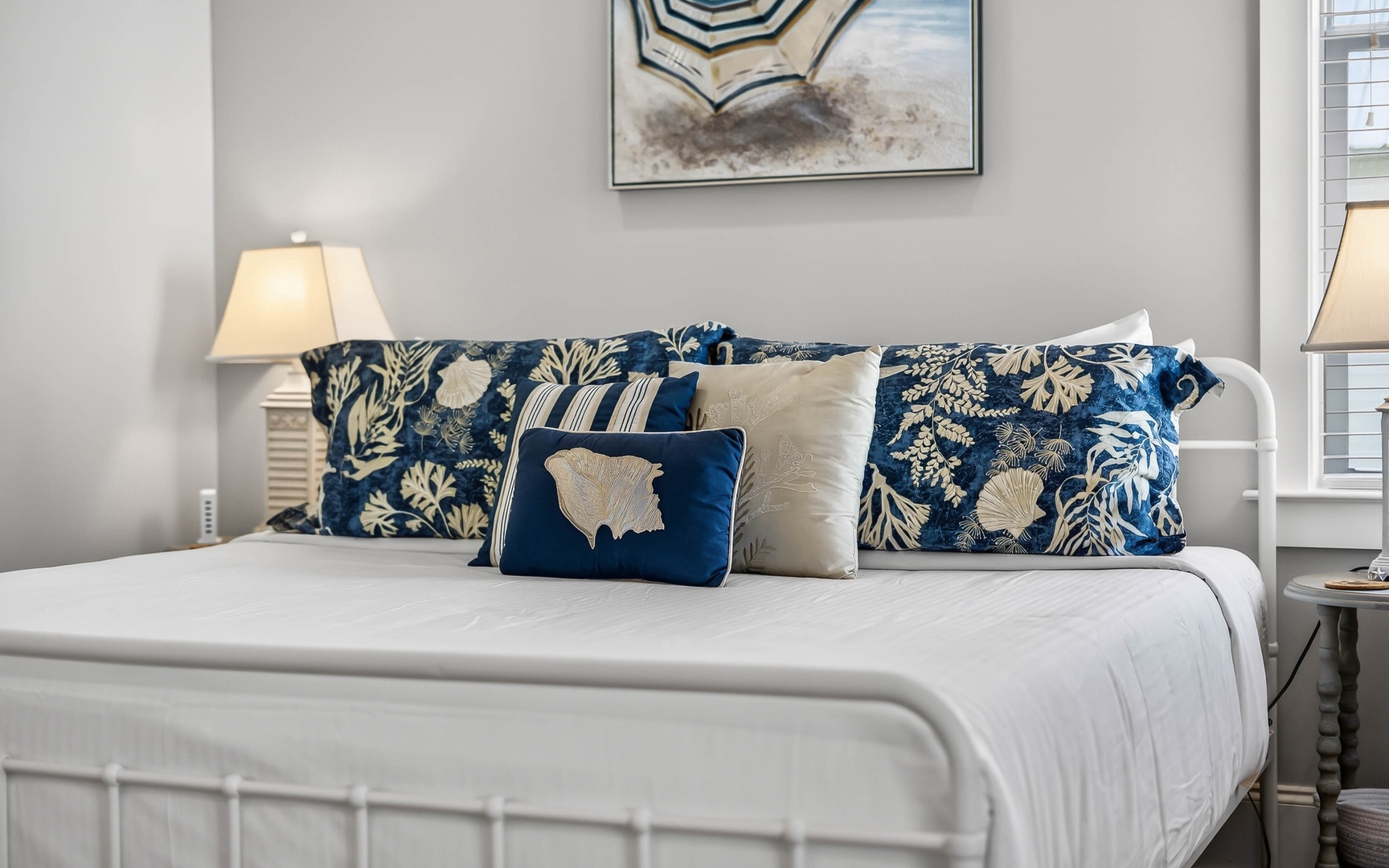 Sink into crisp white linens adorned with coastal-inspired blue pillows, where soft lamplight creates the perfect reading ambiance.