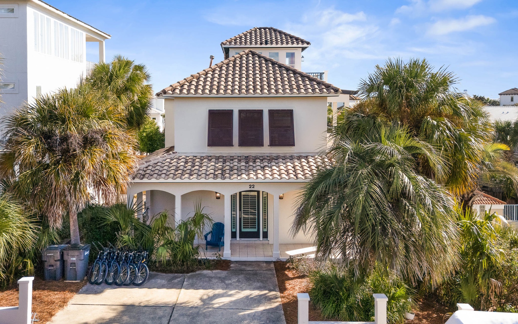Mediterranean-style villa with clay tile roof surrounded by swaying palm trees in a tropical neighborhood setting.