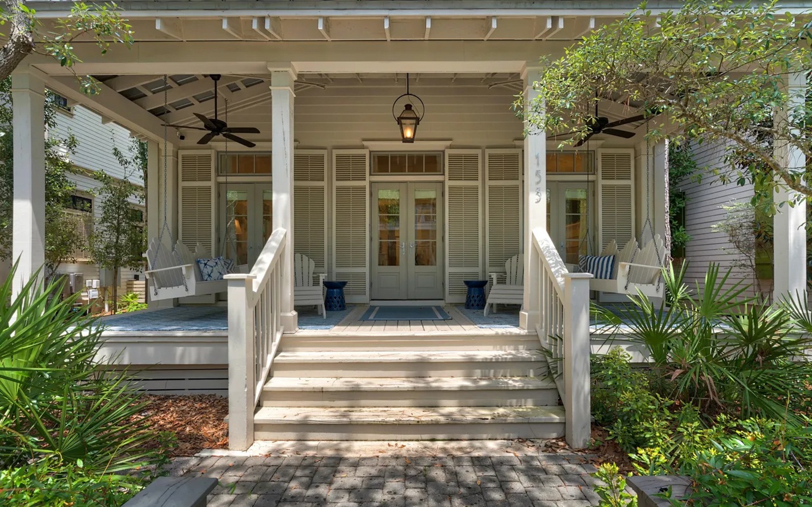 Classic Southern charm welcomes guests with its wraparound porch, elegant columns, and inviting entrance framed by lush tropical landscaping.