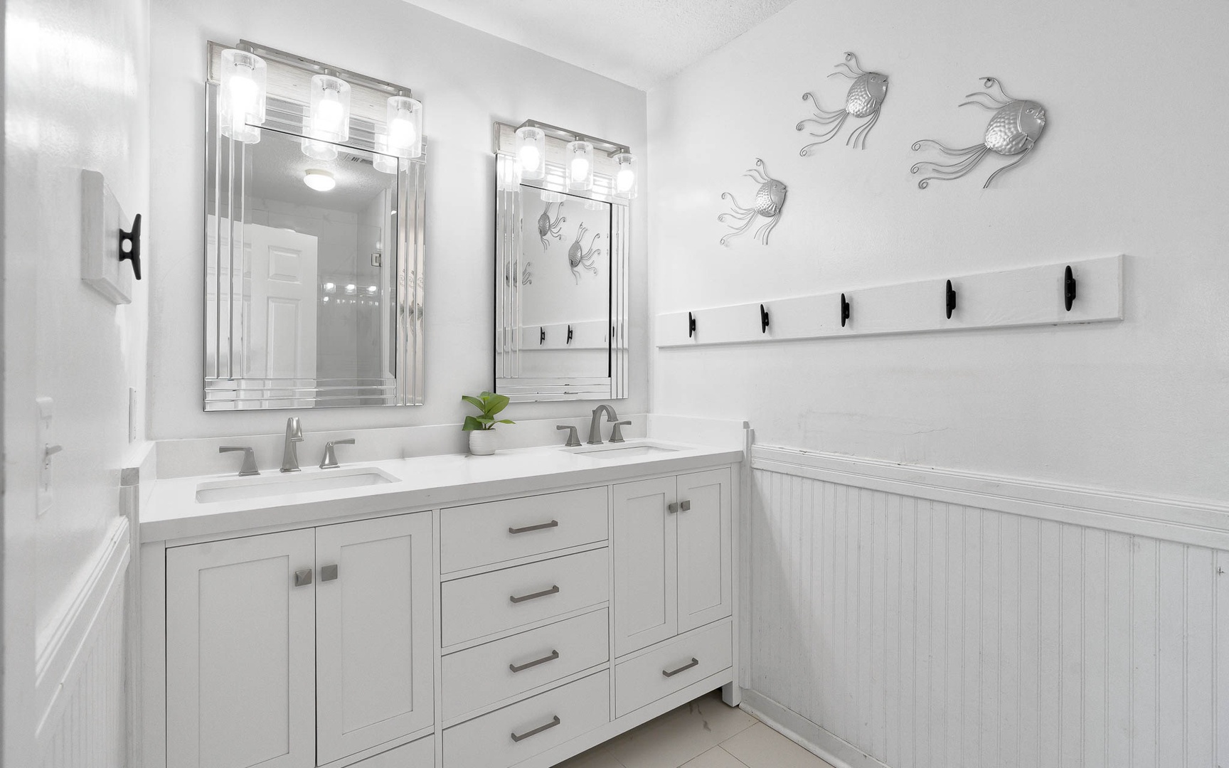 Step into your pristine white bathroom with elegant double vanities, perfect Hollywood-style lighting, and charming coastal fish art.