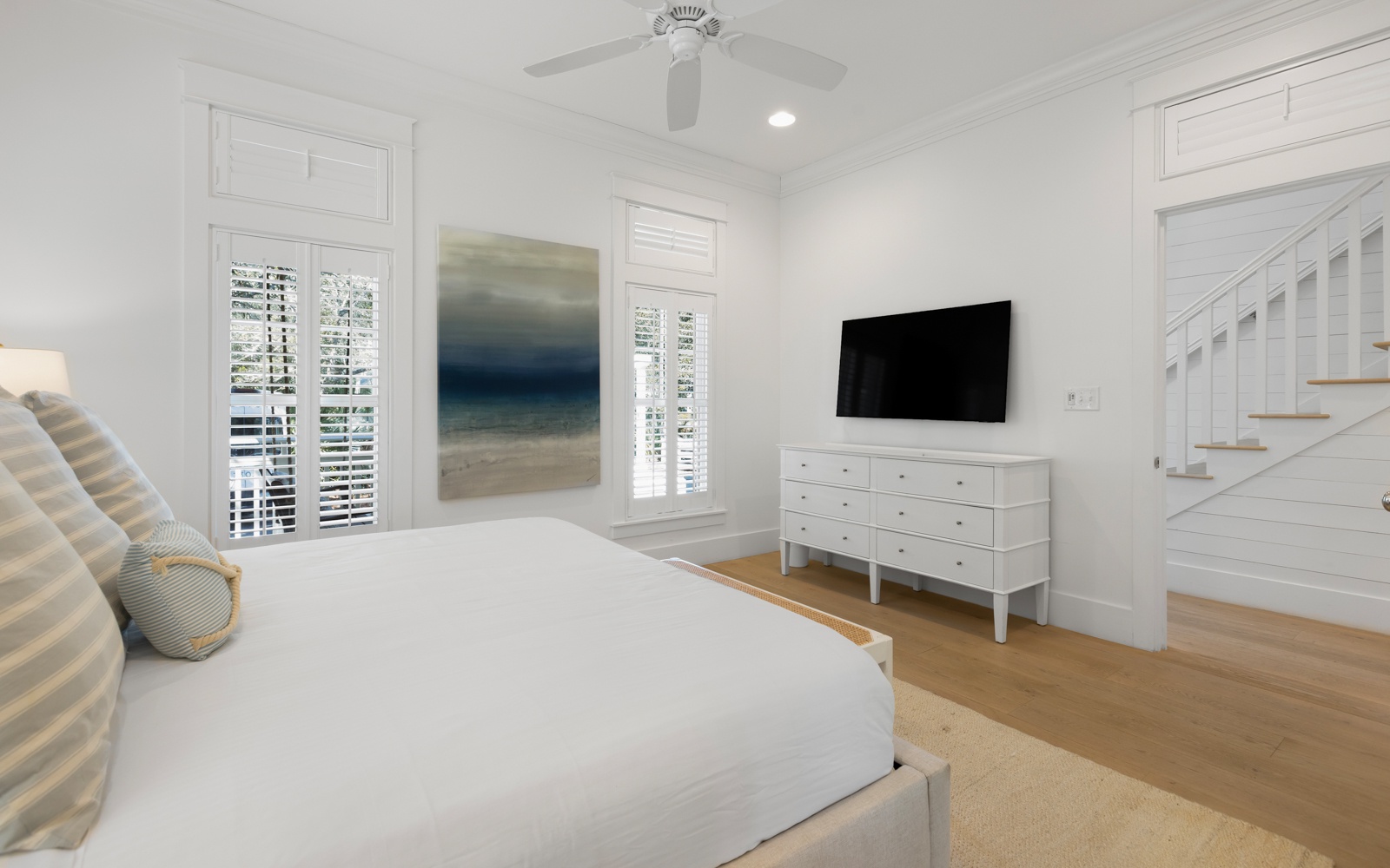 Unwind in your serene white bedroom featuring plush bedding, ocean-inspired artwork, and a wall-mounted TV for cozy nights in.
