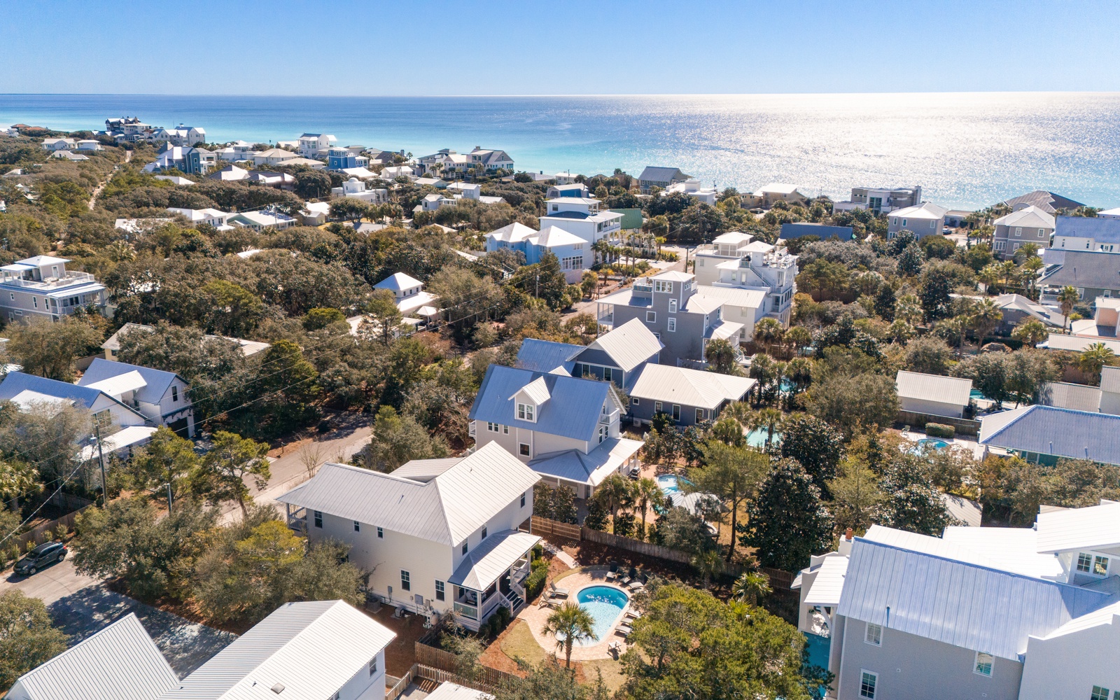 Aerial view of a charming coastal community with white beach houses nestled among lush trees, just steps from pristine turquoise waters.