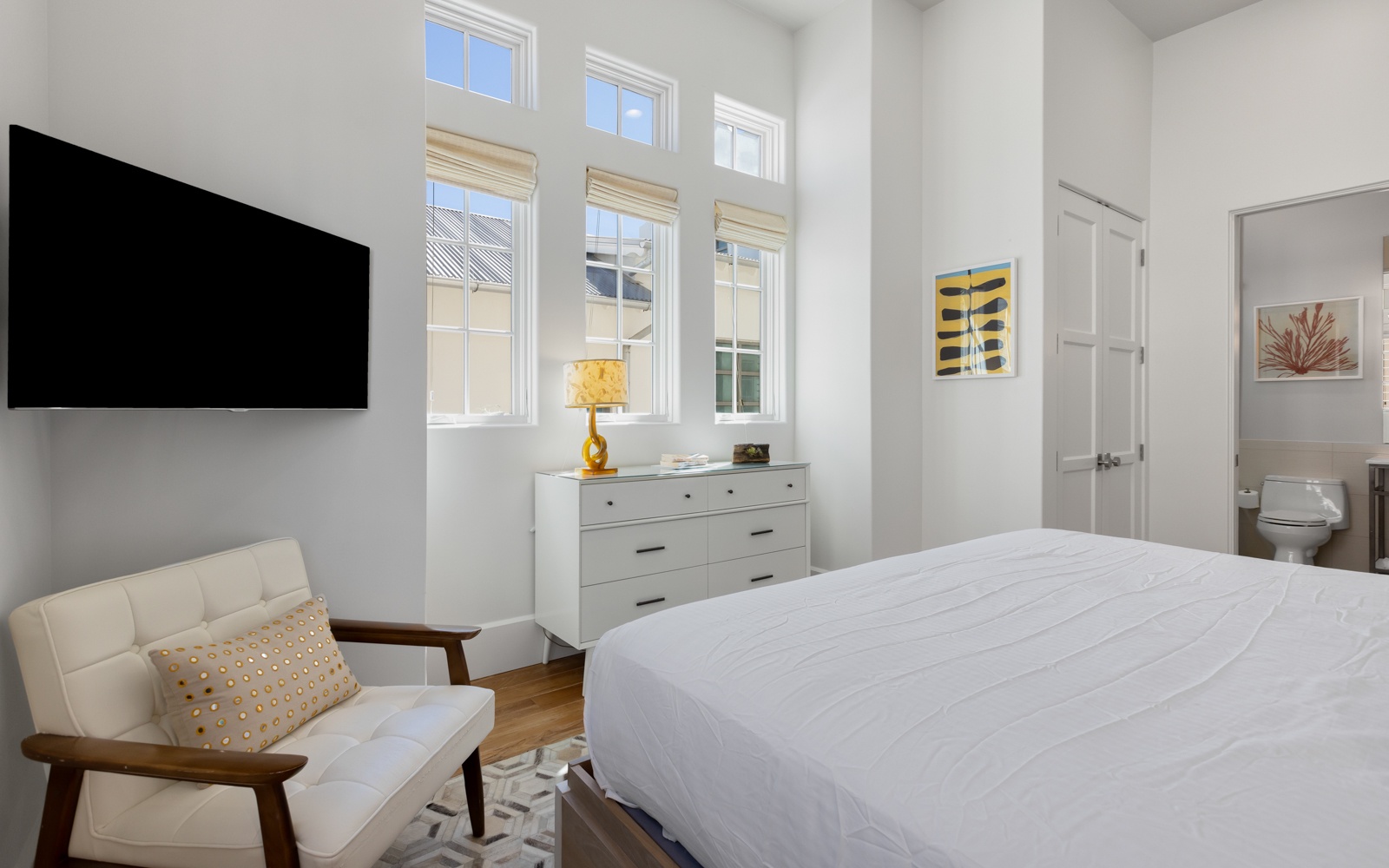 Your bright bedroom features crisp linens, a cozy reading chair, and tall windows flooding the space with natural light.