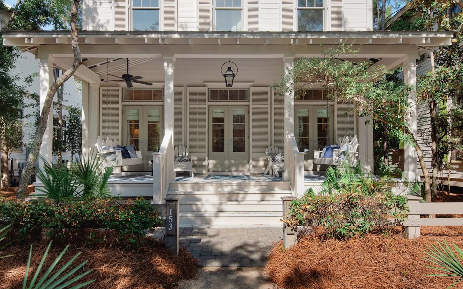 Charming white cottage with wraparound porch and comfortable seating surrounded by lush landscaping and mature trees.