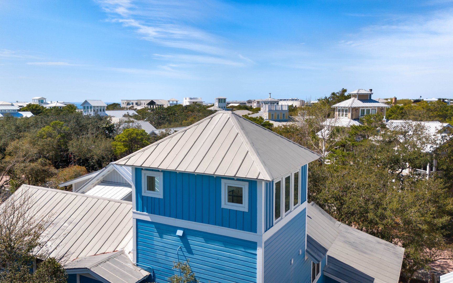Bright blue coastal home nestled among trees in a charming neighborhood, with metal roofing and surrounding vacation properties visible.