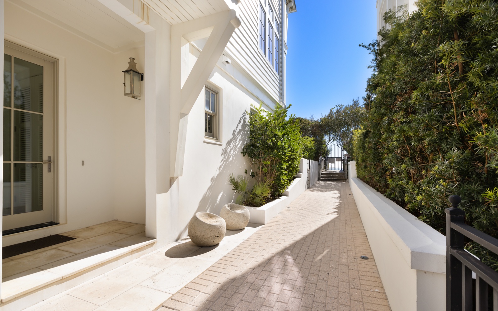A pristine white exterior walkway with decorative planters leads toward the entrance, surrounded by lush greenery under bright blue skies.