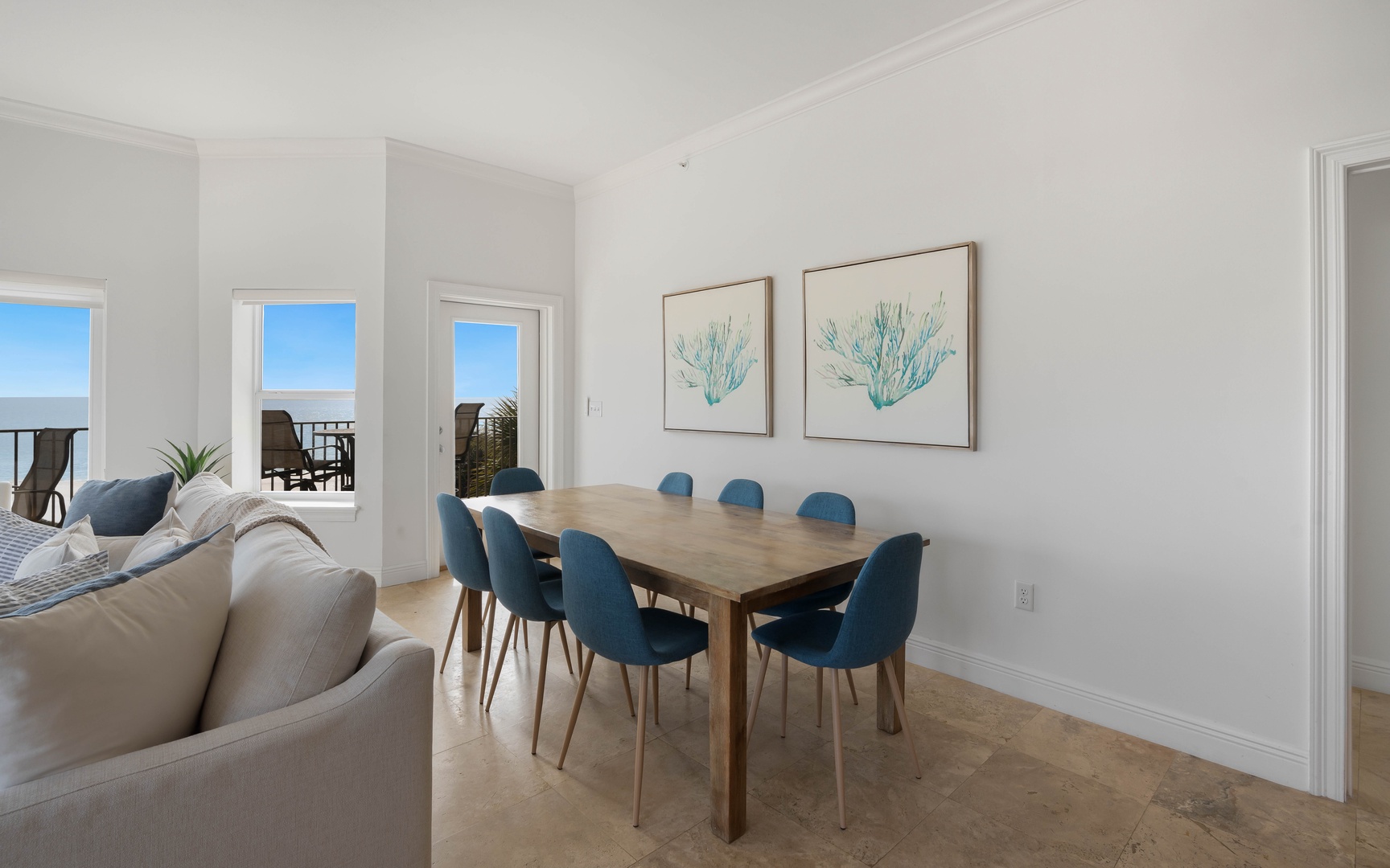 Your bright dining space opens to stunning ocean views through floor-to-ceiling windows, where coastal artwork complements the serene blue palette.