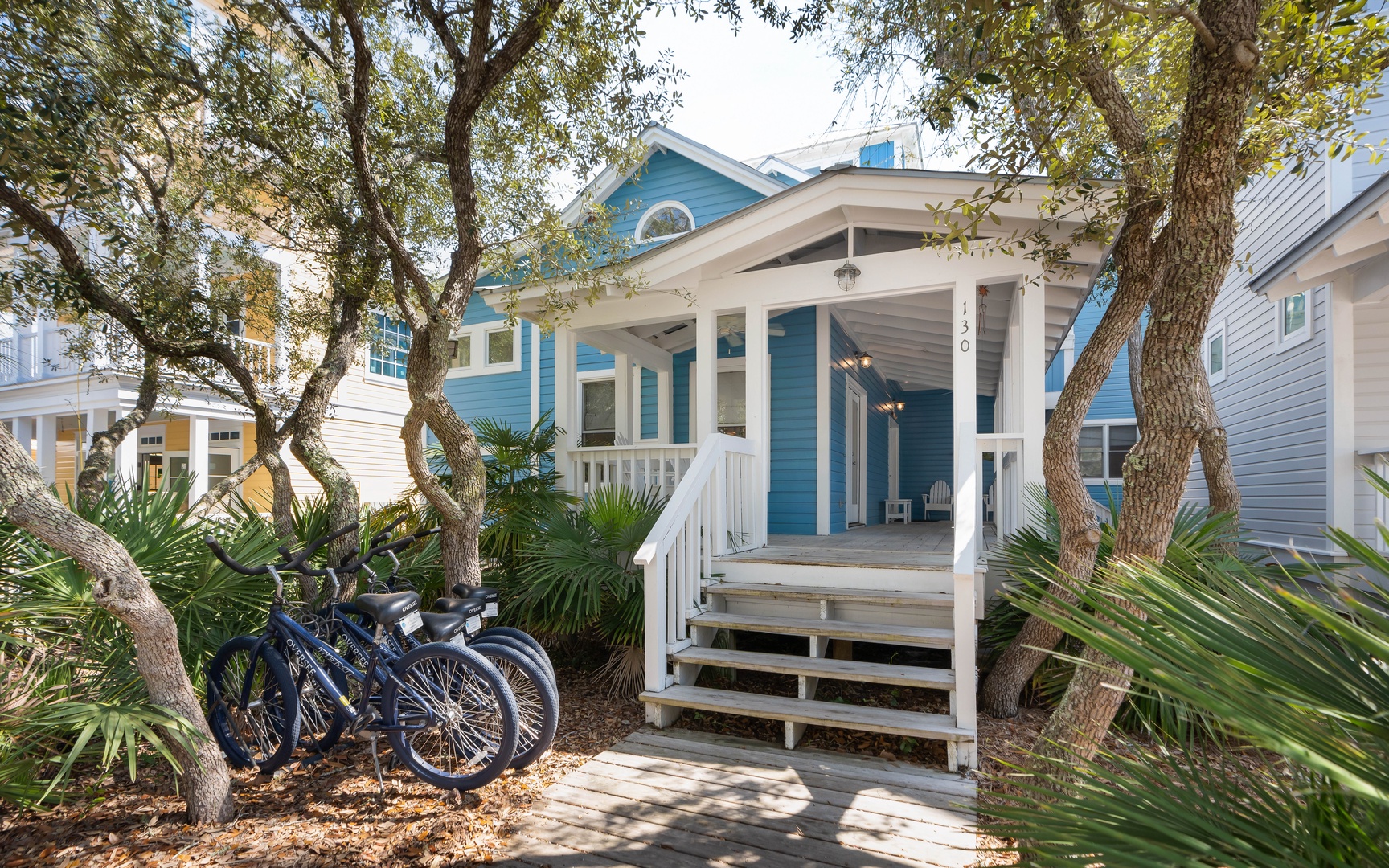 Charming coastal cottage with welcoming front porch, surrounded by mature trees and tropical landscaping in a peaceful neighborhood setting.