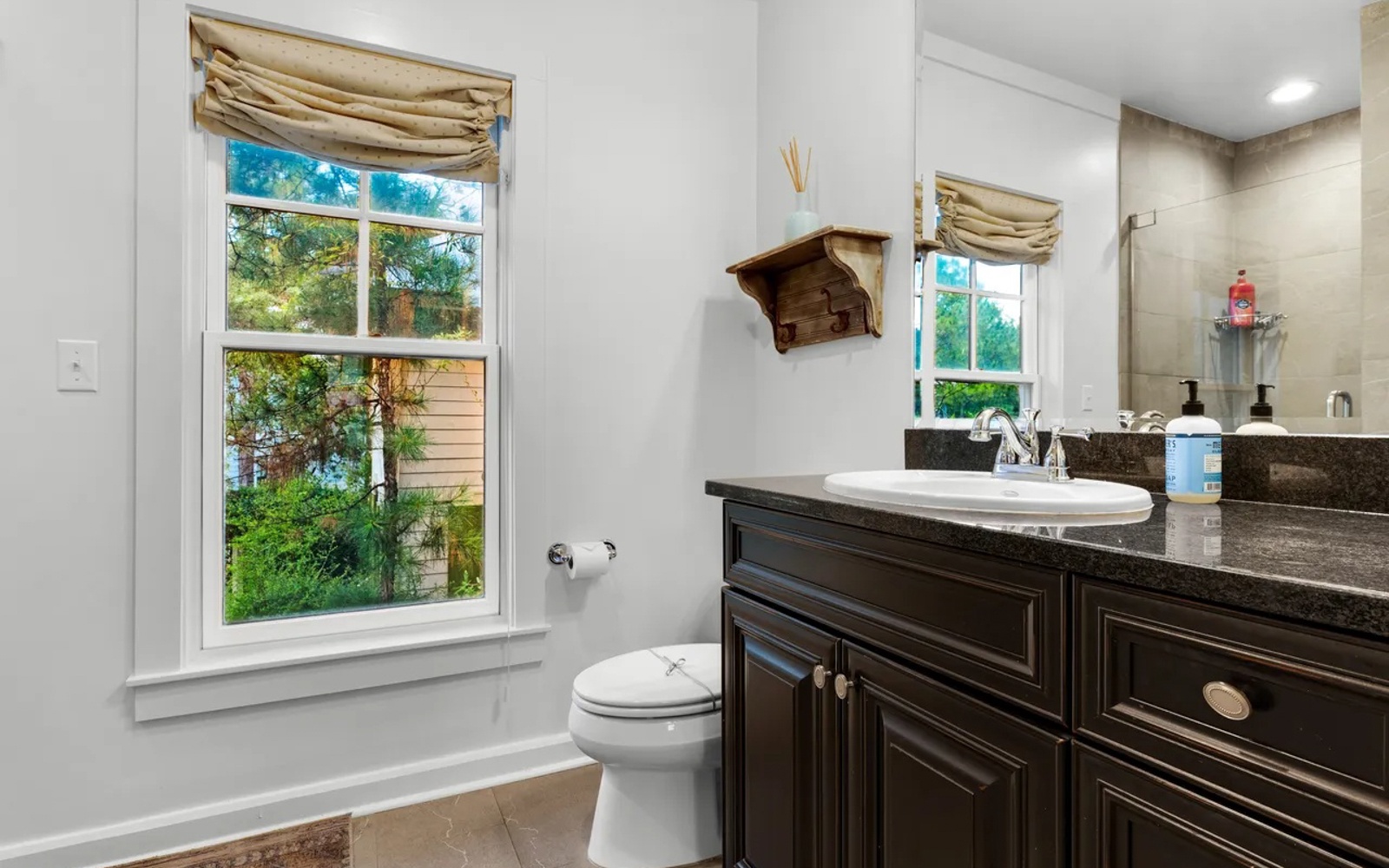 Refresh in your elegant bathroom featuring dark granite counters, garden views through charming windows, and thoughtful touches like natural linen shades.