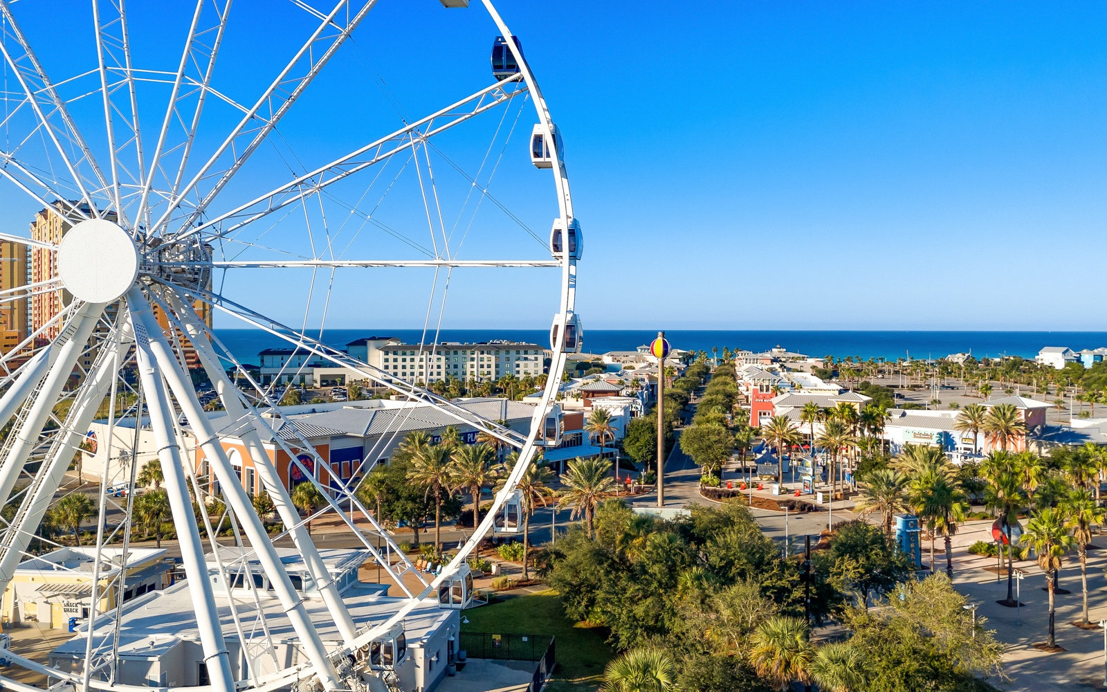 White ferris wheel overlooks a coastal city with palm trees, colorful buildings, and ocean views stretching to the horizon.