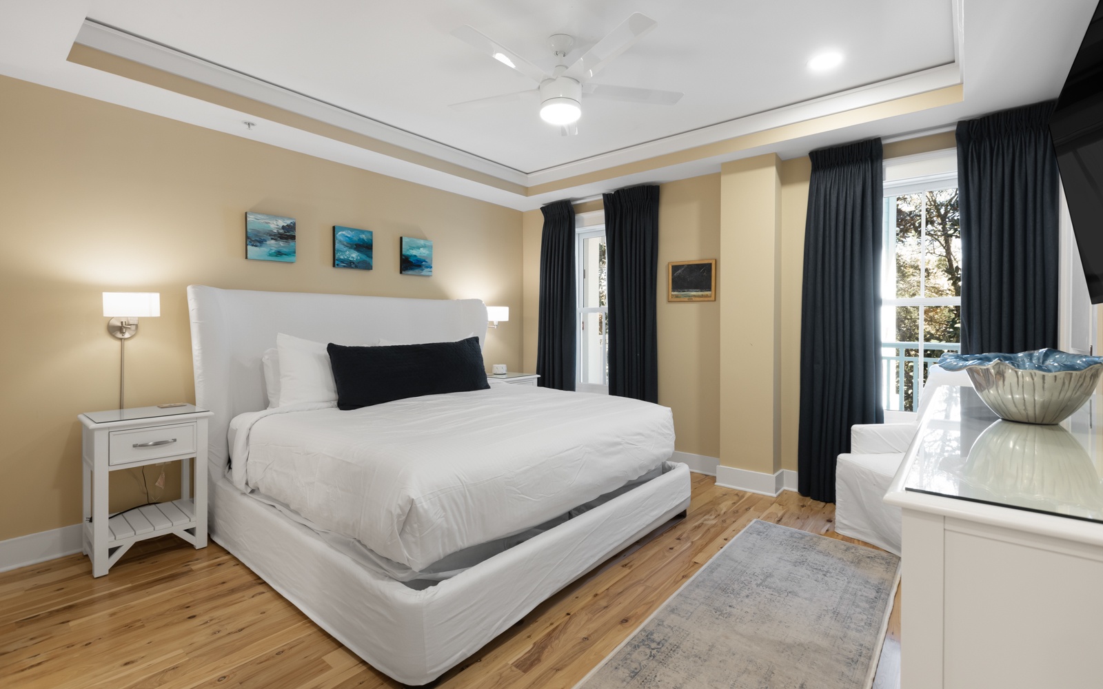 Unwind in your peaceful bedroom featuring a plush white bed, warm wood floors, and serene ocean artwork above your headboard.