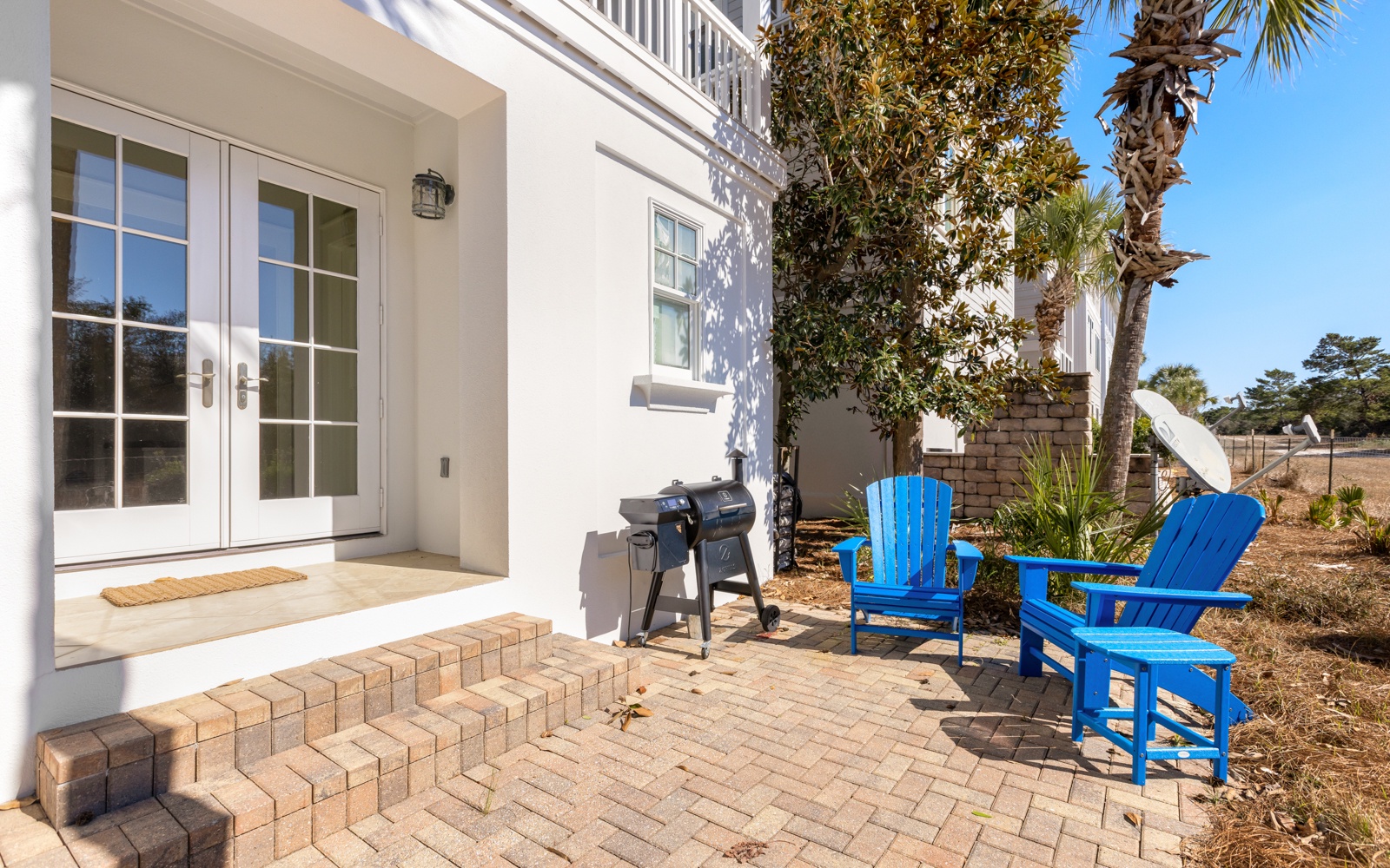 Your private patio invites relaxation with bright blue Adirondack chairs, BBQ grill, and tropical landscaping under sunny skies.
