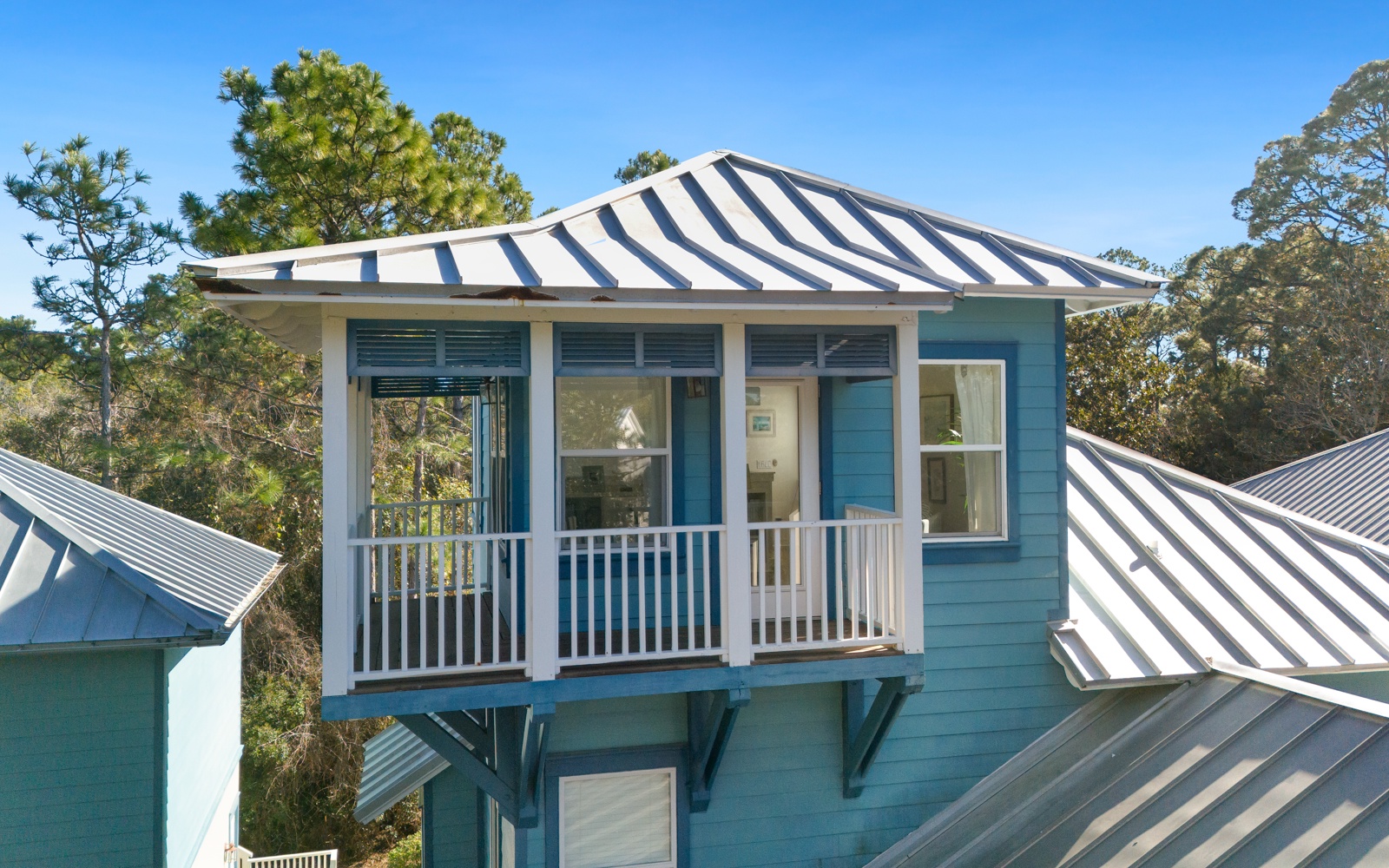 Charming coastal cottage with cheerful turquoise siding and striped awning overlooking lush treetops.