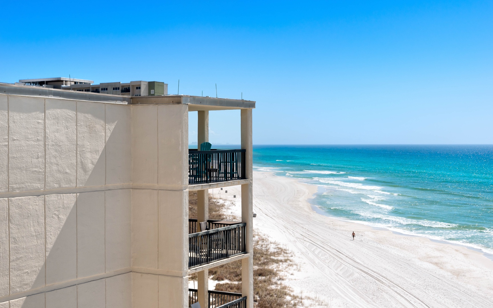 Pristine white sand beach stretches along turquoise waters with modern beachfront property featuring multi-level balconies overlooking the stunning Gulf Coast.