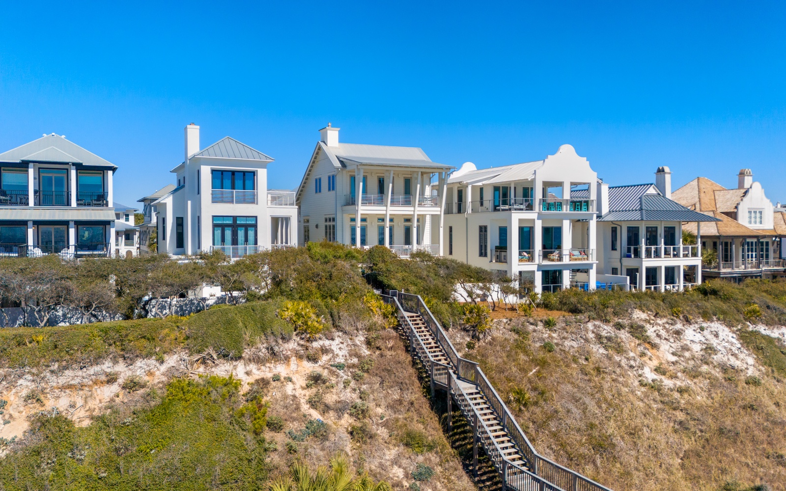 Stunning beachfront homes line the coast with natural dune vegetation and wooden boardwalk access under brilliant blue skies.