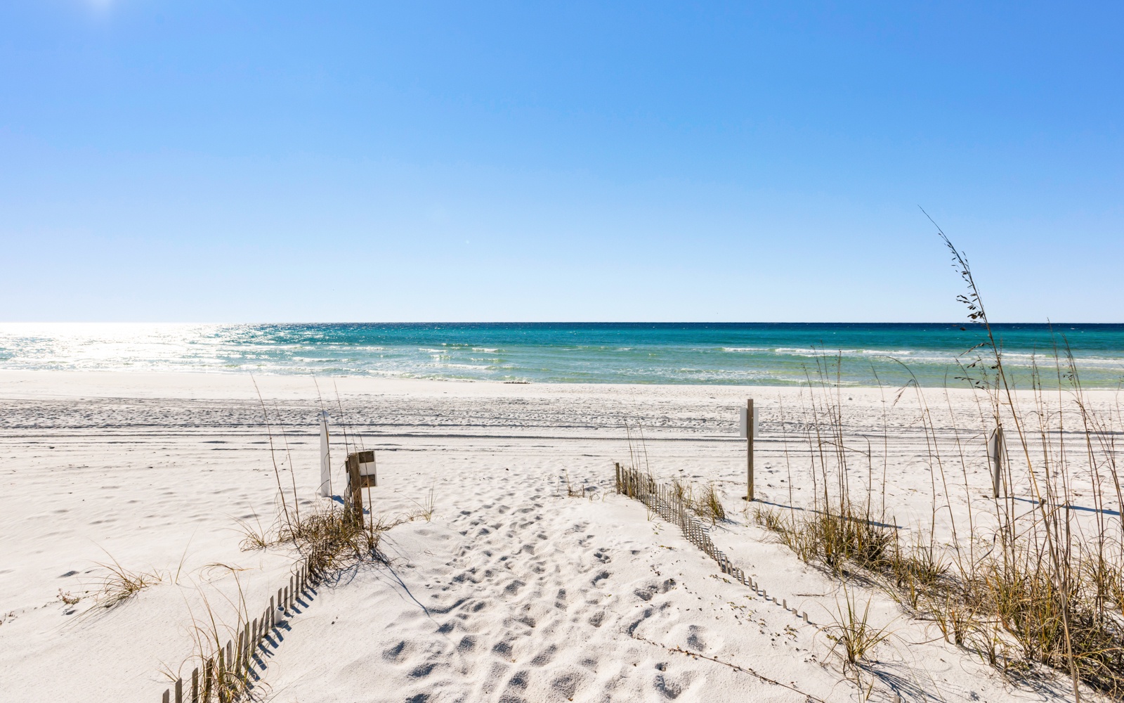 Pristine white sand beach with turquoise waters and dunes, featuring a scenic boardwalk path to the shoreline.