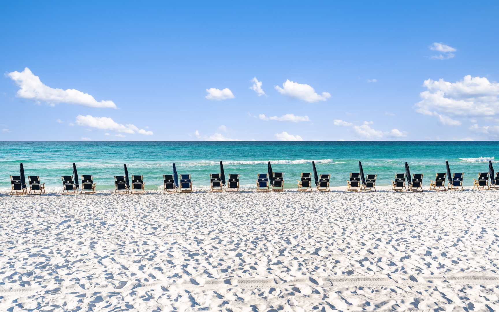 Pristine white sand beach with rows of umbrellas and chairs overlooking turquoise waters under blue skies.