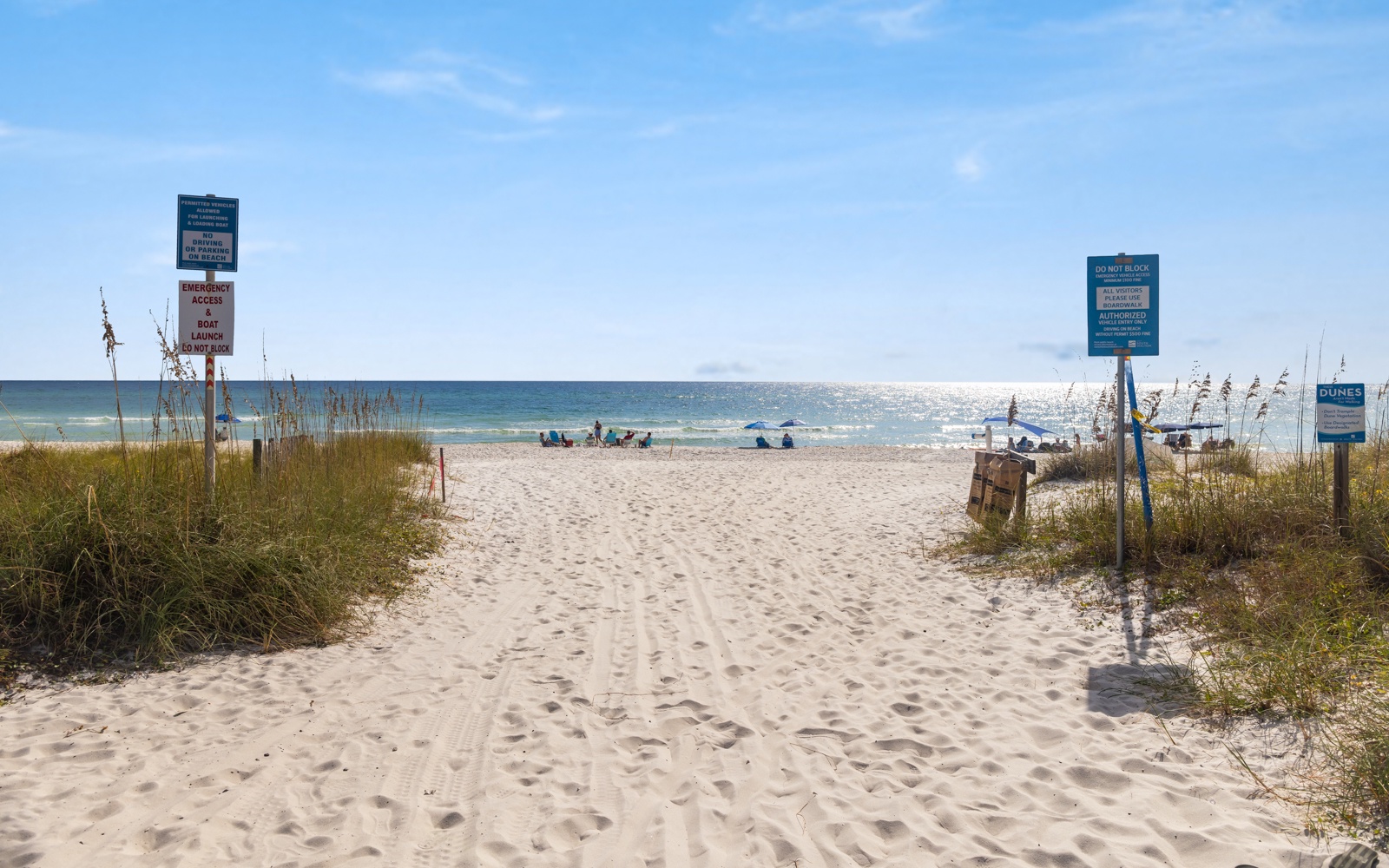 Beach access pathway leads through coastal dunes to pristine sandy shores with clear blue waters and gentle waves.