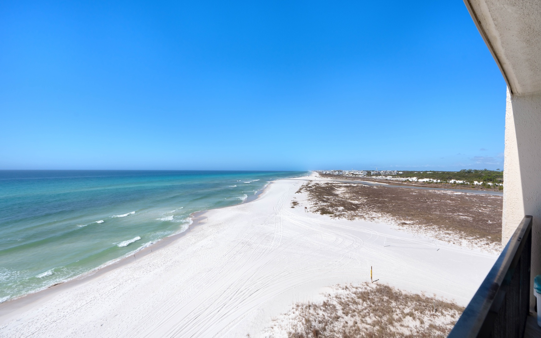 Pristine white sand beach stretches endlessly along turquoise waters under clear blue skies, captured from an elevated oceanfront balcony.