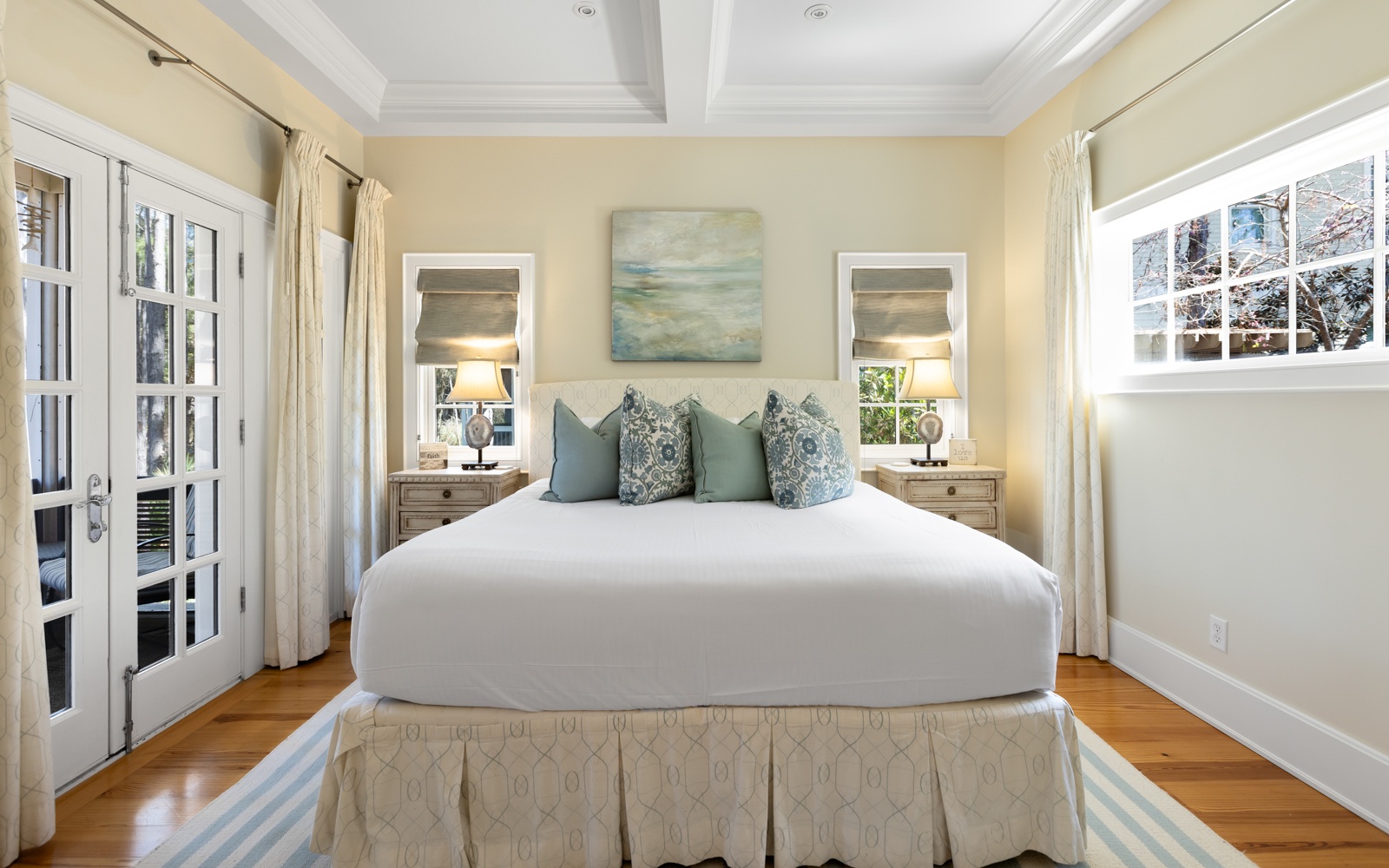 Drift into peaceful sleep on this plush bed, surrounded by warm coastal colors and soft natural light from French doors.