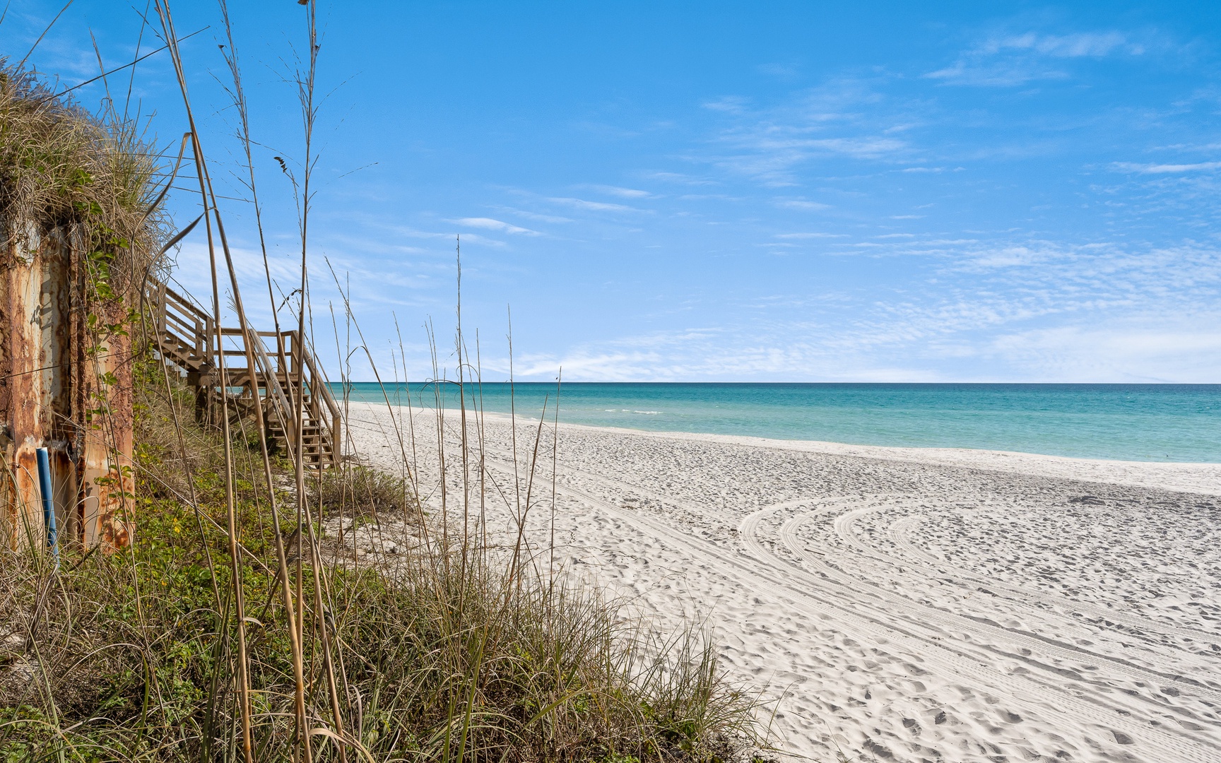 Pristine white sand beach with wooden boardwalk access through natural dunes, offering stunning turquoise waters and endless coastal views.