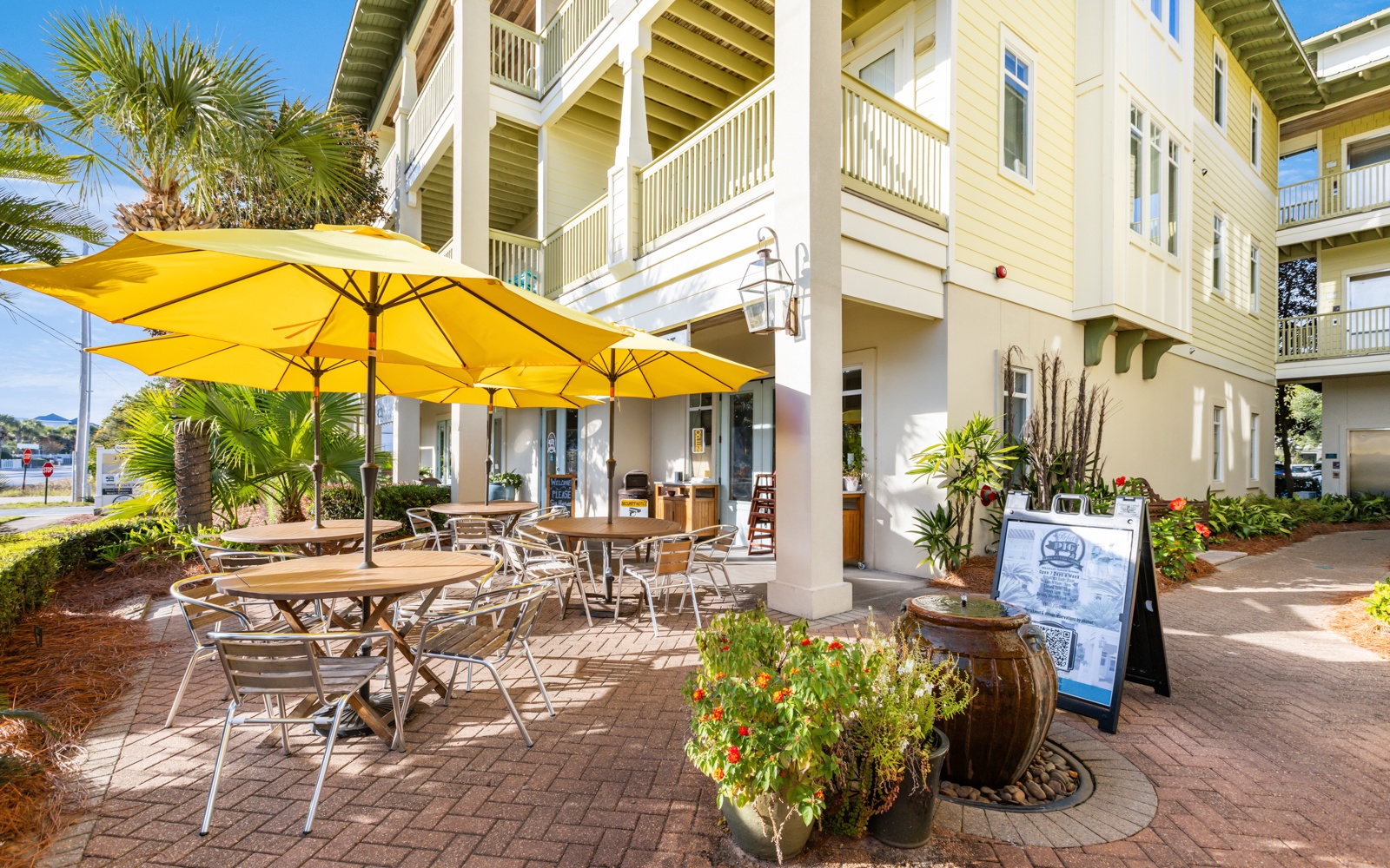 Charming beachside restaurant with outdoor dining under sunny yellow umbrellas, surrounded by tropical landscaping and coastal architecture.