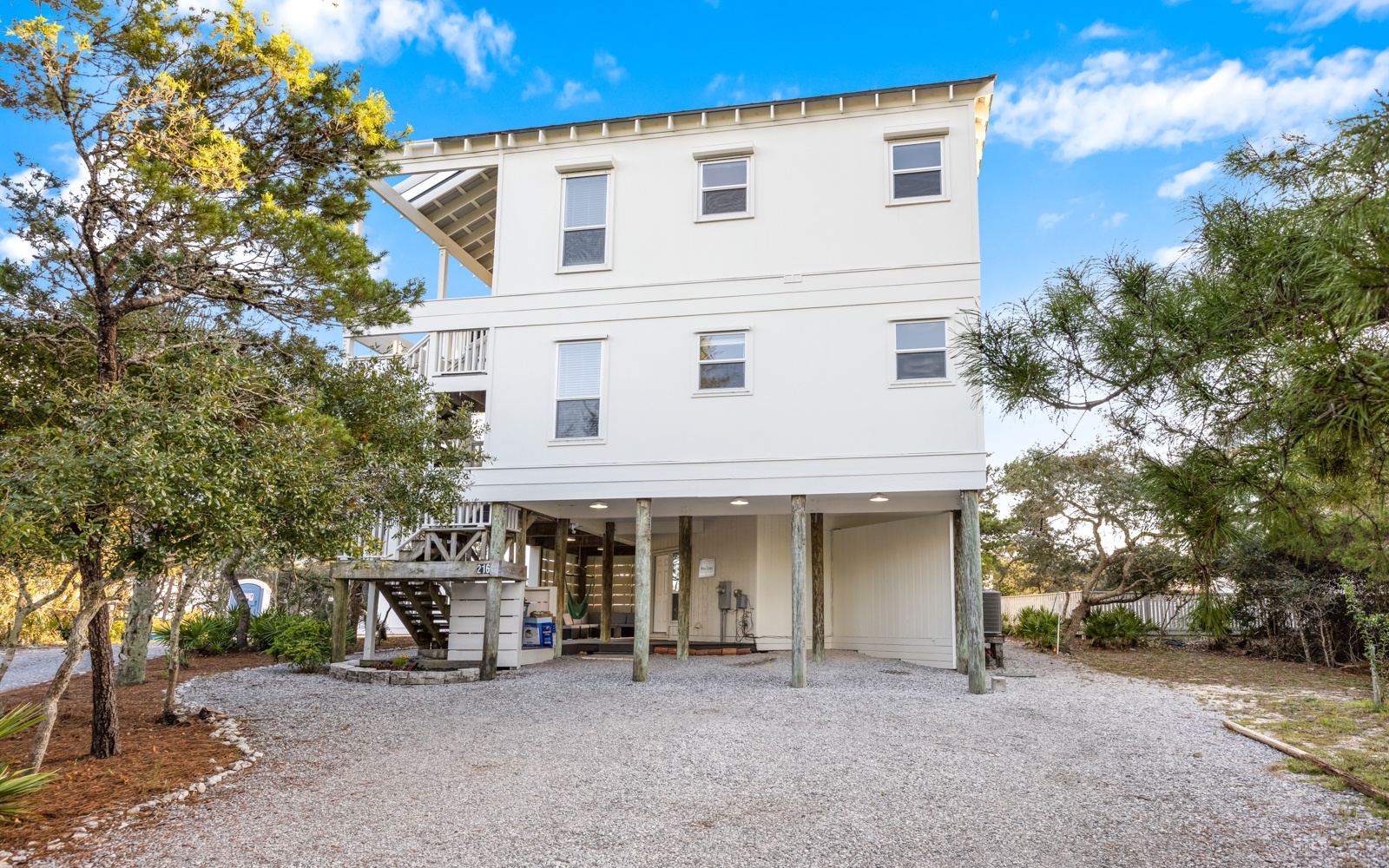 Elevated beach house with covered parking and outdoor stairs, surrounded by coastal trees under bright blue skies.