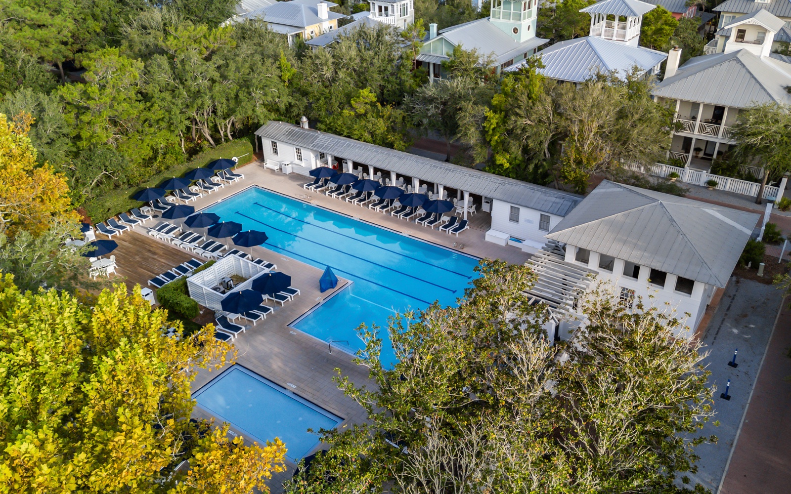 Aerial view showcases the property's impressive pool complex with multiple swimming areas, surrounded by lush coastal trees and charming neighborhood architecture.
