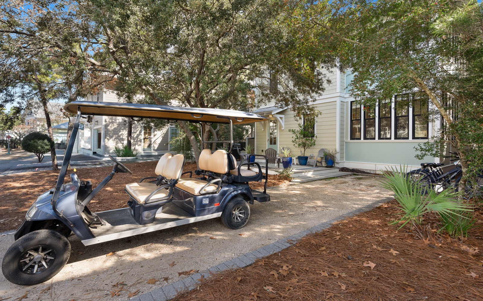 Golf cart parked outside modern beach house with covered porch surrounded by mature trees in coastal setting.